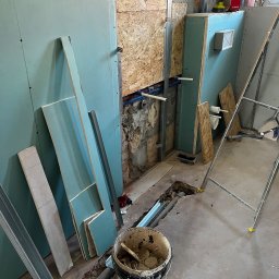 Builder Atta - Montaż płyt gipsowo-kartonowych w łazience, widoczne profile, płyty GK, instalacja wodna, drabina i wiadro z zaprawą. Etap prac wykończeniowych w pomieszczeniu.