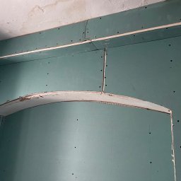 Builder Atta - Niebiesko-zielone płyty karton-gips tworzące łukowate przejście w ścianie. Widoczne łączenia płyt i ślady po wkrętach. Surowy stan wykończenia wnętrza.