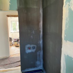 Builder Atta - Przygotowanie ścian podłoża pod płytki w łazience. Na ścianach widoczna szara hydroizolacja, wystające rury zasilające. W tle widoczne narzędzia i materiały budowlane.