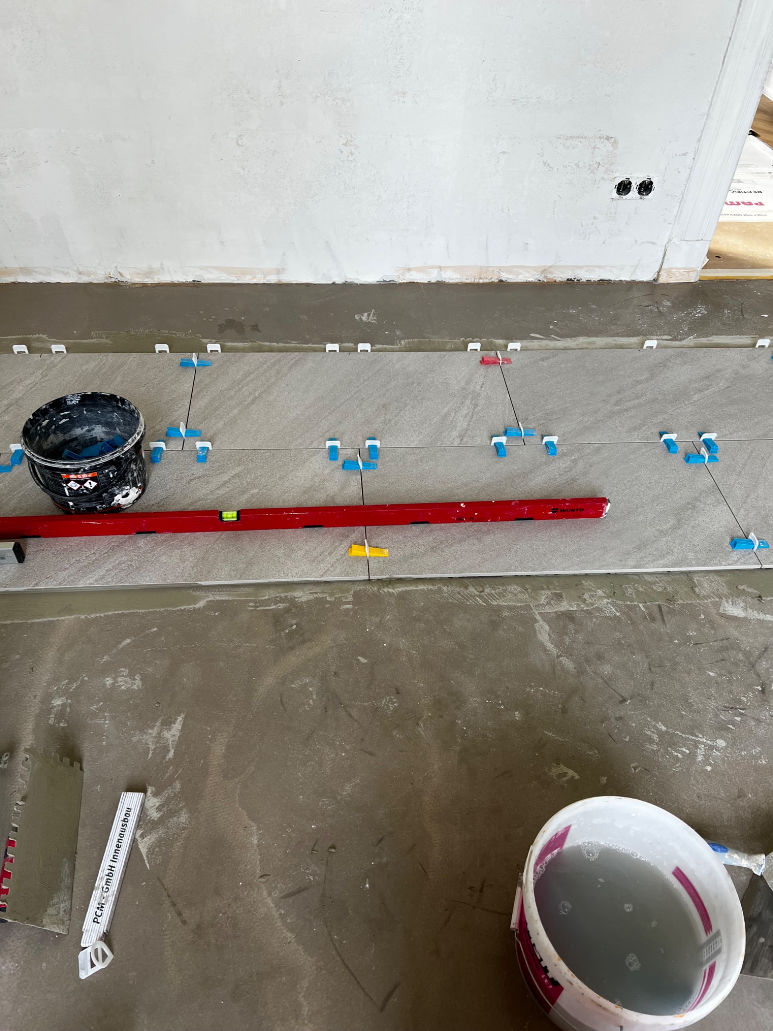 Układanie szarych płytek podłogowych z użyciem poziomicy, krzyżyków dystansowych i dwóch wiader z zaprawą na surowym podłożu.