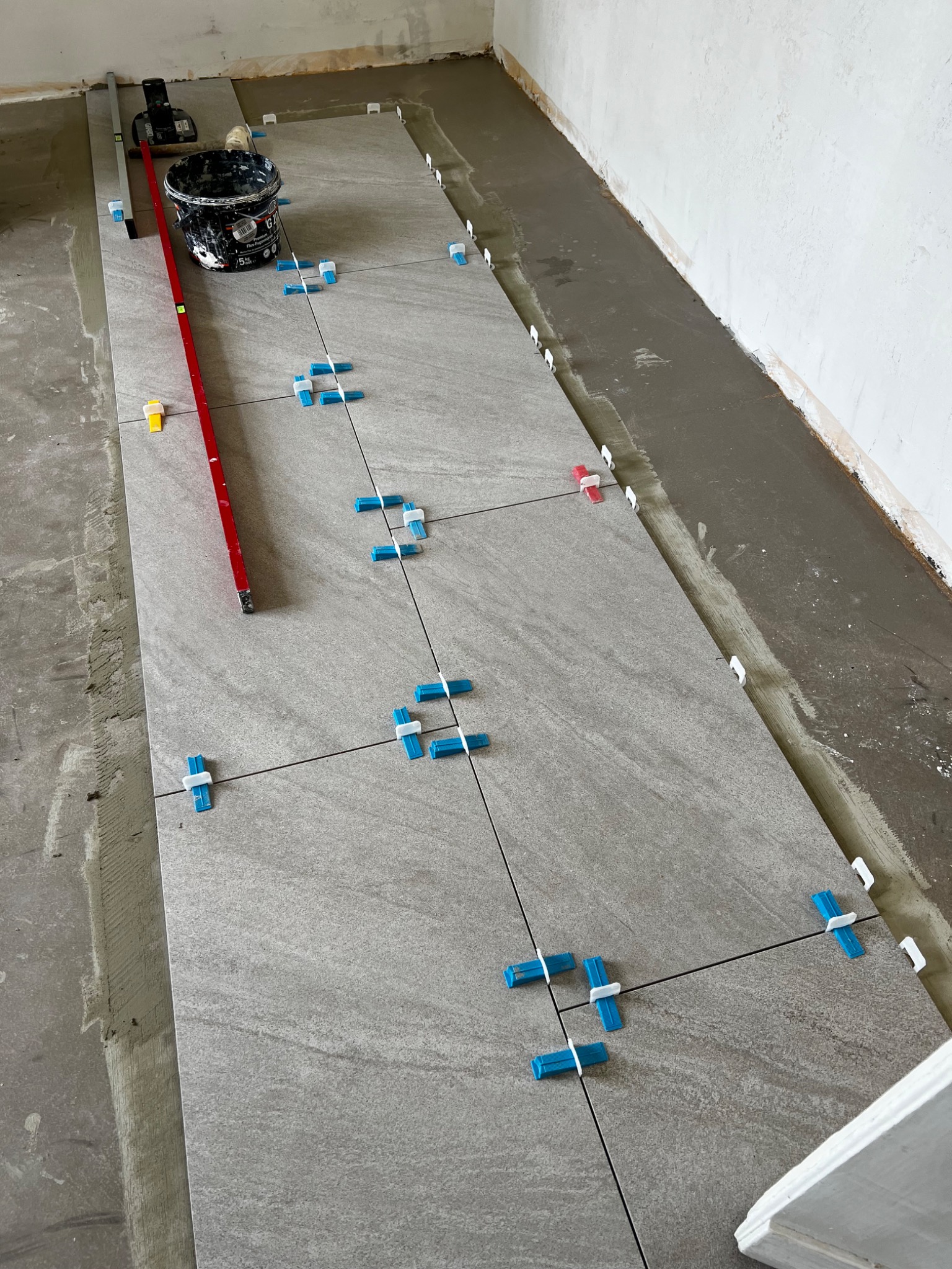 Ułożone płytki podłogowe w trakcie montażu, z niebieskimi i białymi krzyżykami dystansowymi, poziomica w tle, widoczny klej i narzędzia.