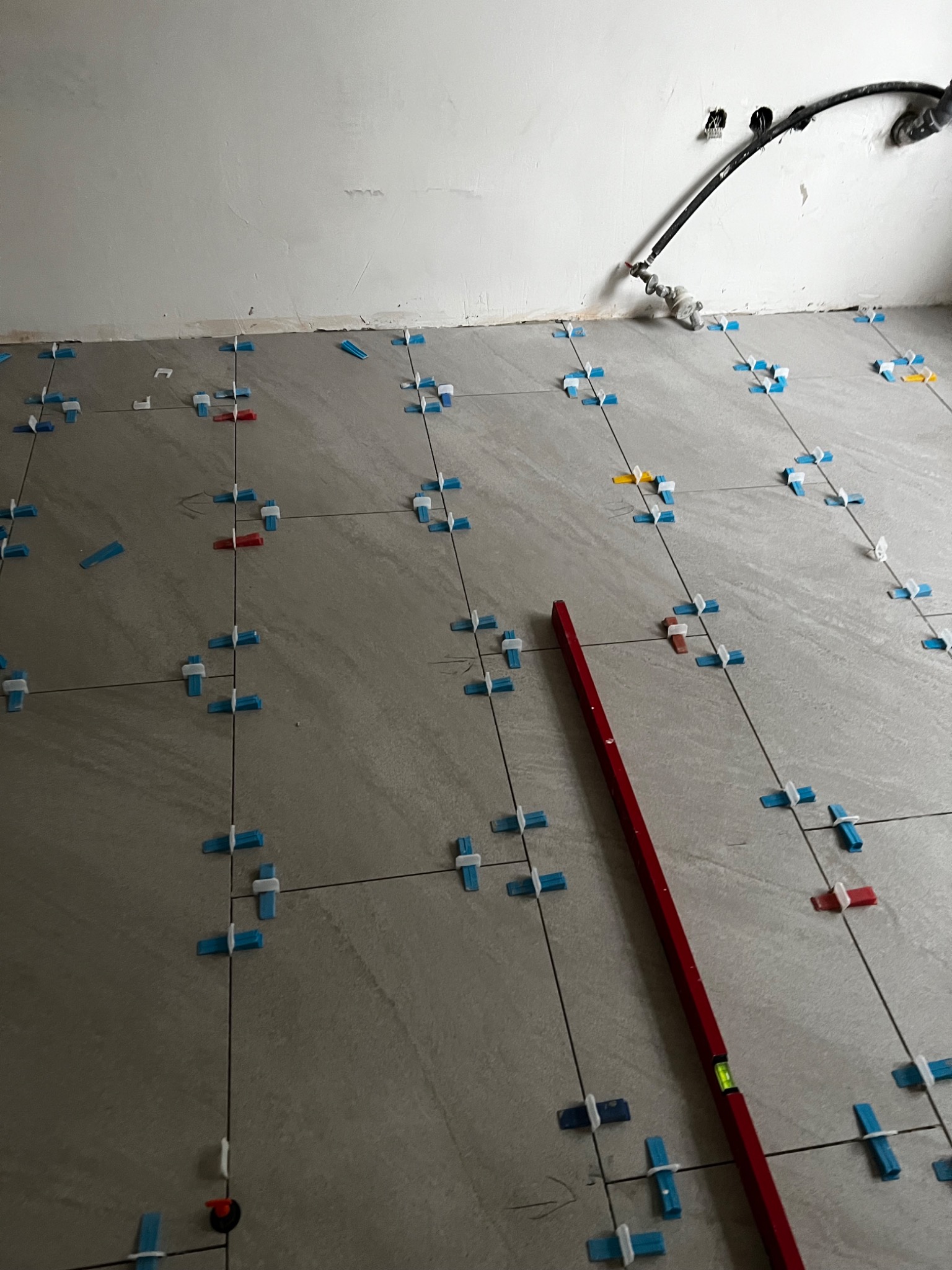Układanie szarych płytek podłogowych z użyciem niebieskich i czerwonych krzyżyków dystansowych, poziomica wzdłuż fugi, surowy stan ściany w tle.