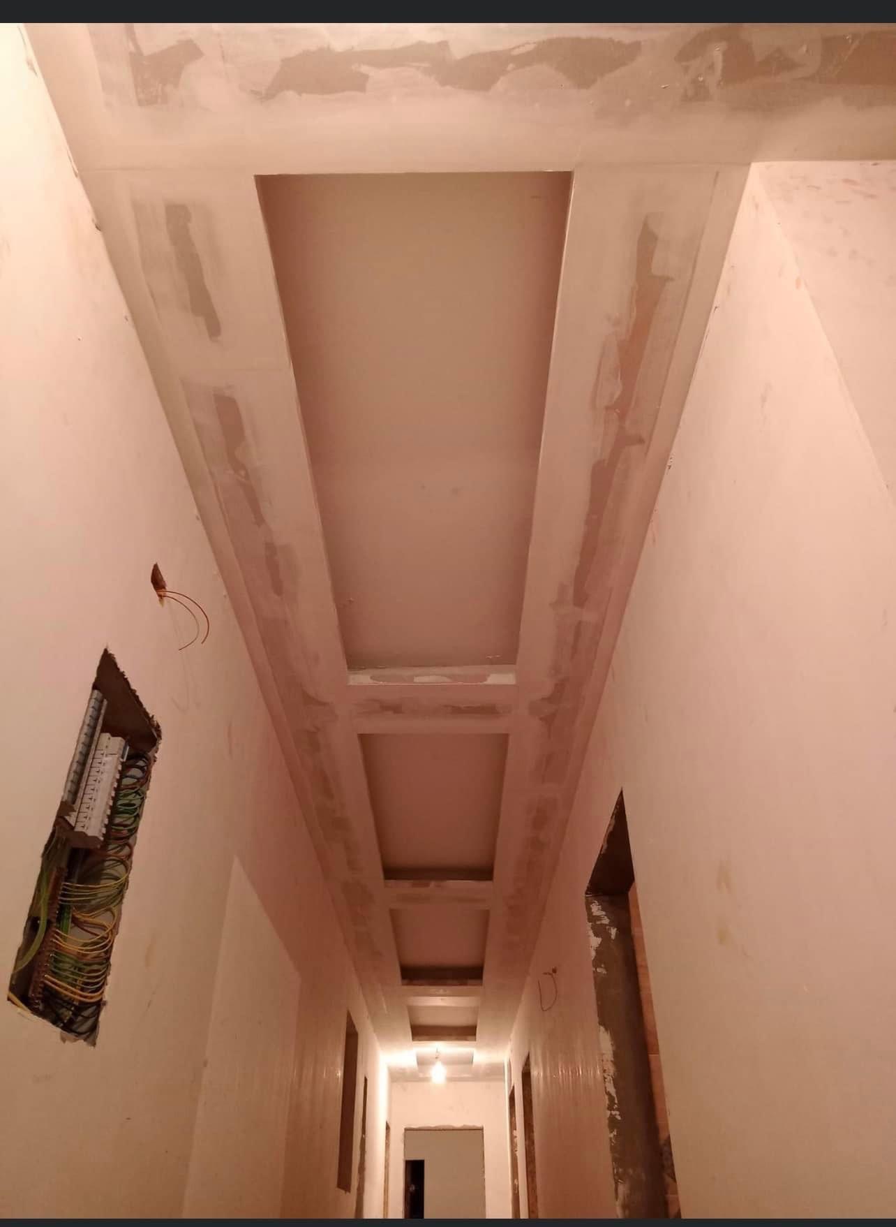 Korytarz w trakcie prac wykończeniowych, widoczne podwieszane sufity z wnękami oraz otwarty panel elektryczny z okablowaniem na ścianie. Ściany przygotowane pod malowanie.