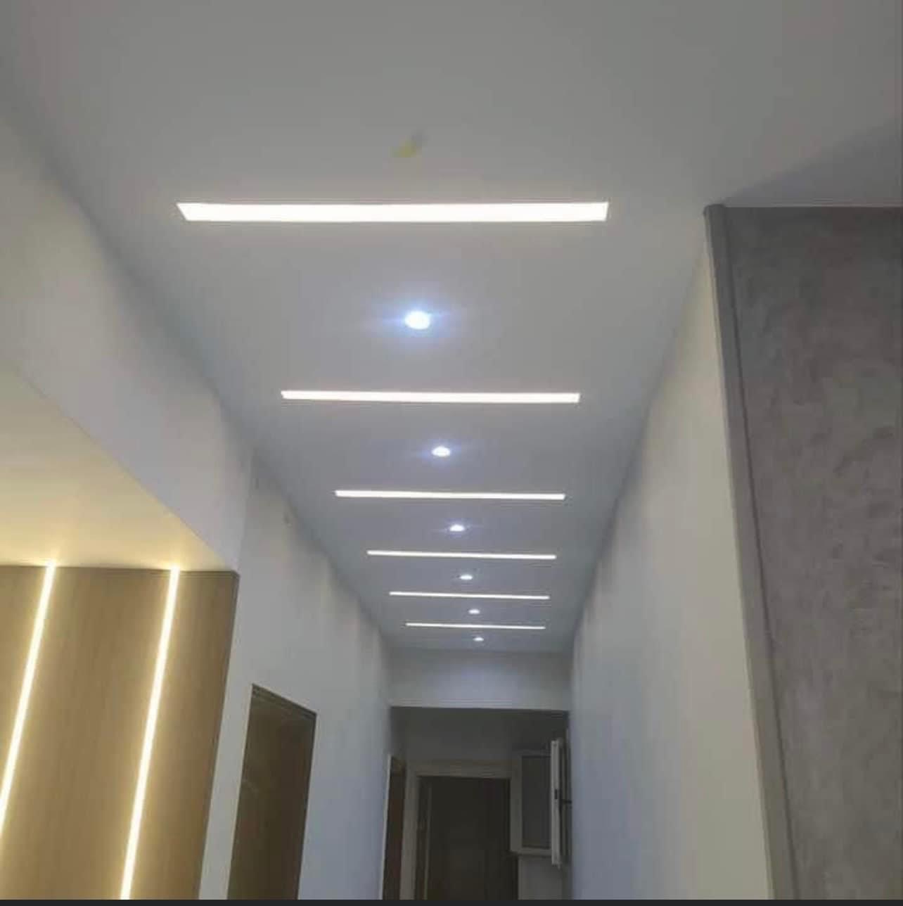 Długi korytarz z nowoczesnym oświetleniem sufitowym w postaci wpuszczonych pasków LED i punktowych lamp, ściany wykończone w różnych odcieniach szarości, widoczny fragment drewnianej okładziny...