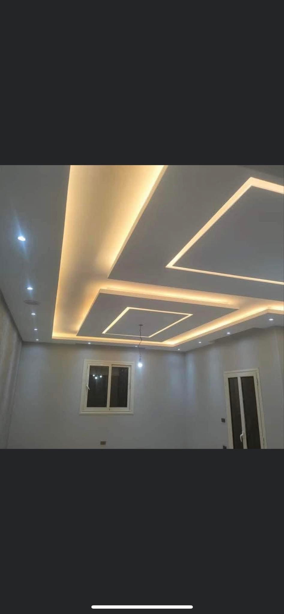 Nowoczesny sufit podwieszany z oświetleniem LED w kształcie kwadratów, świeżo wykończone ściany w jasnych odcieniach, okno i drzwi widoczne w tle.