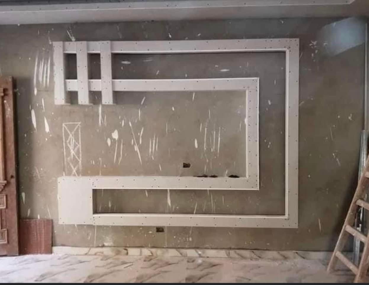 Niedokończona konstrukcja z płyt kartonowo-gipsowych na szarej ścianie, widoczne ślady szpachlowania, drabina oparta o ścianę, marmurowa podłoga w tle.