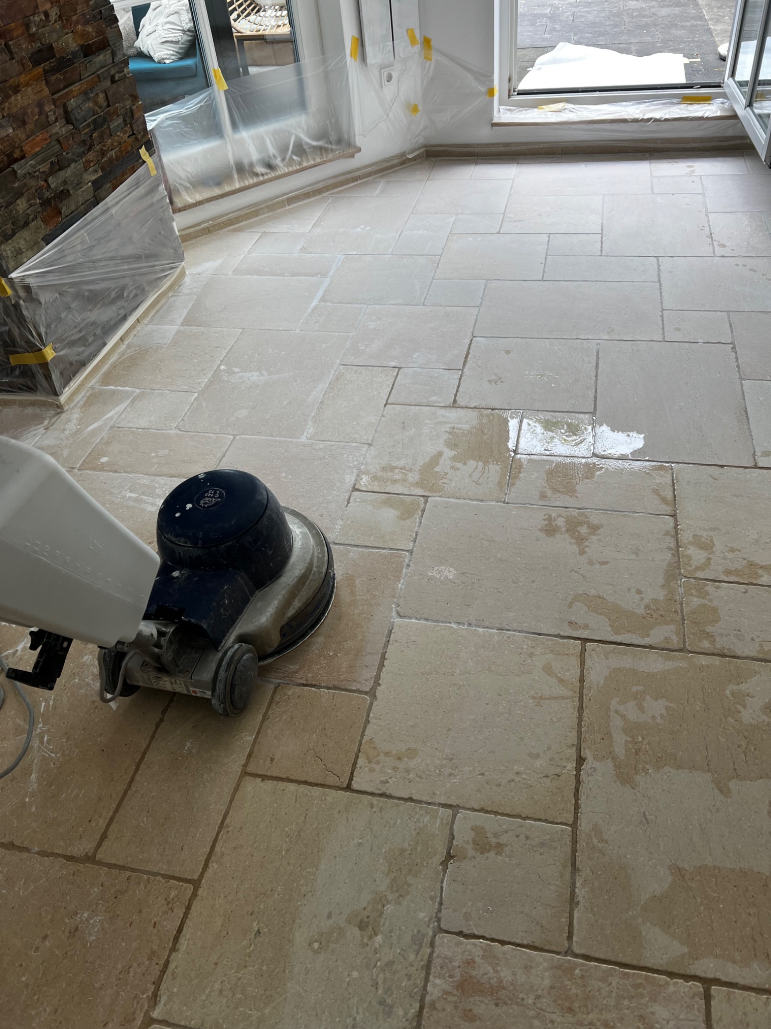 Maszyna do czyszczenia kamiennej podłogi w pomieszczeniu z zabezpieczonymi folią oknami i kamienną ścianą.