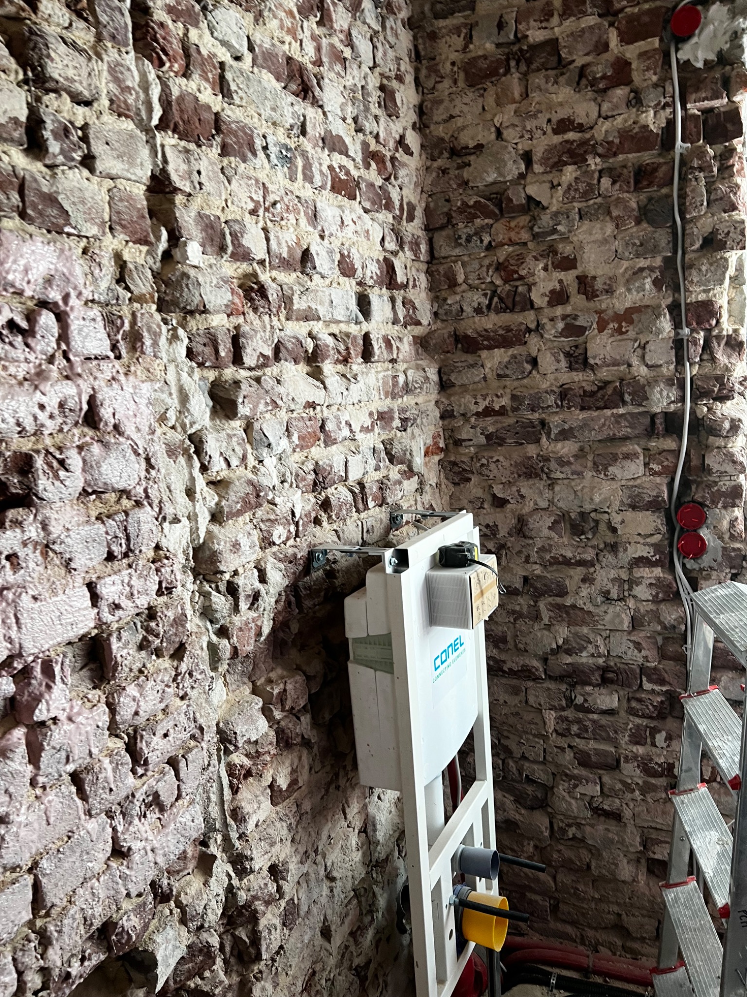 Montaż stelaża podtynkowego WC na tle surowej ściany z cegły, widoczne przewody elektryczne i drabina.