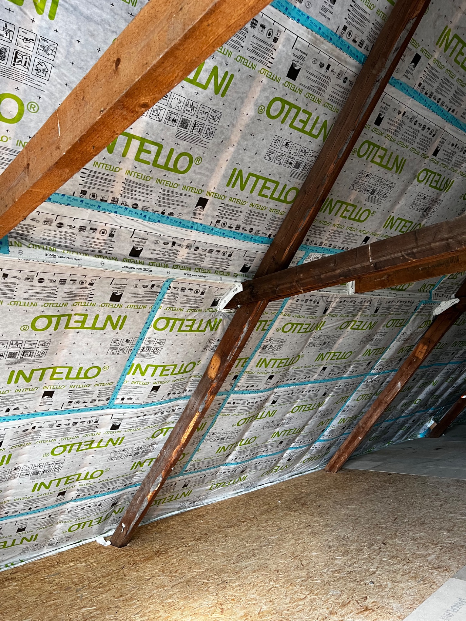 Poddasze z widoczną izolacją termiczną marki Intelio, zamontowaną między krokwiami, zabezpieczoną taśmą uszczelniającą, widoczne drewniane elementy konstrukcji dachu i fragment podłogi z płyty OSB.