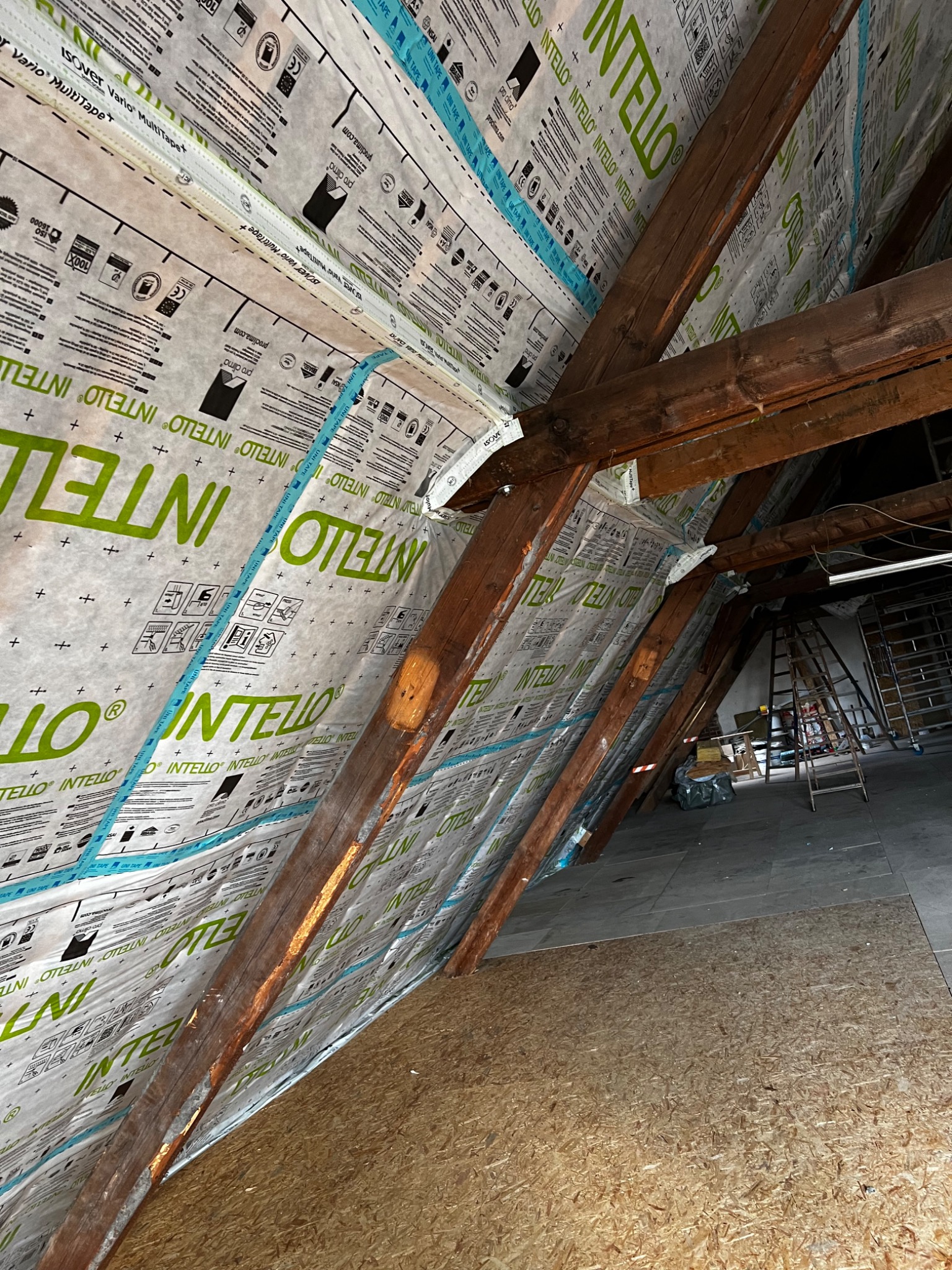 Montaż membrany paroizolacyjnej Intello na drewnianej konstrukcji poddasza, widoczne krokwie i płyty OSB, taśma uszczelniająca na łączeniach, w tle drabiny i elementy wyposażenia.