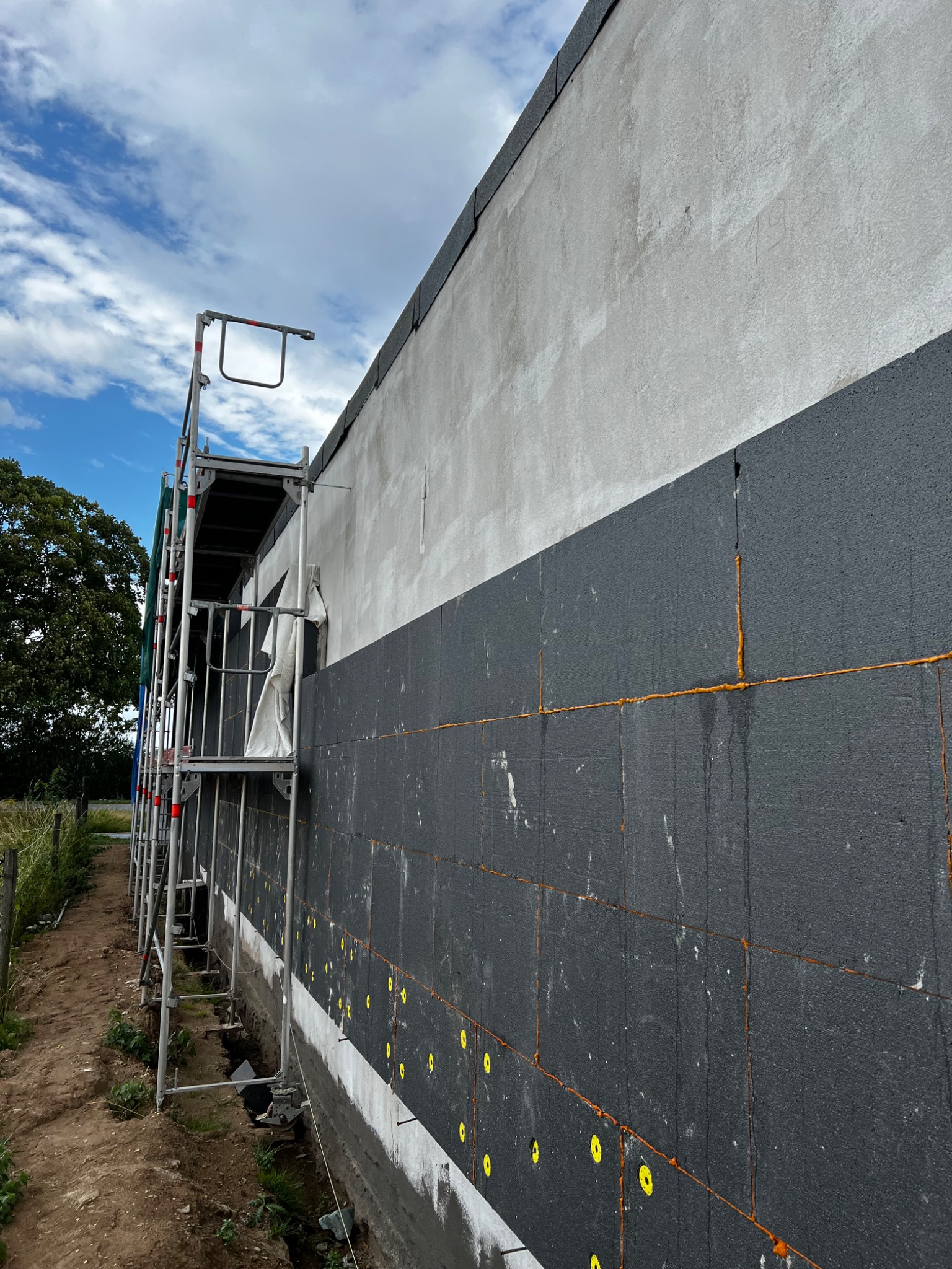 Ocieplanie ściany zewnętrznej budynku styropianem grafitowym, widoczne żółte kołki mocujące, szpary wypełnione pianką montażową, rusztowanie przy ścianie.