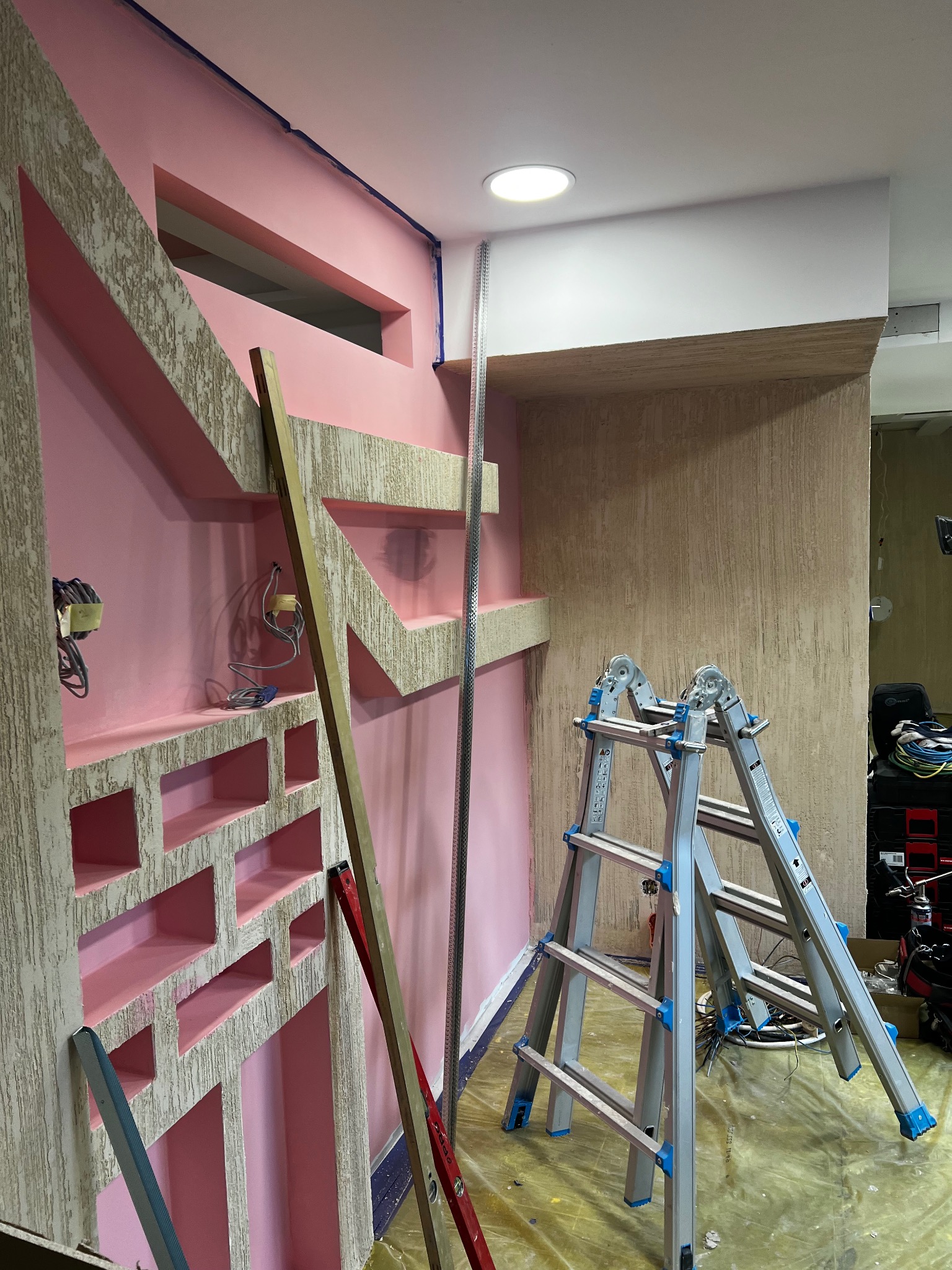 Ściana z dekoracyjnymi elementami w odcieniach różu i beżu, w trakcie prac wykończeniowych. Widoczne drabiny, listwy i przewody elektryczne.
