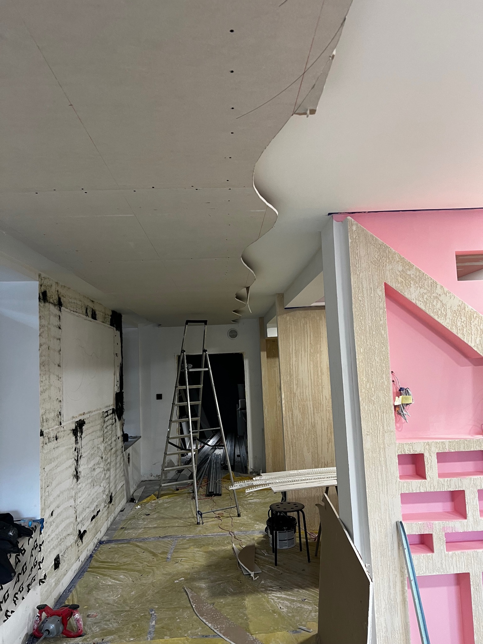 Wnętrze w trakcie remontu: widoczna surowa ściana, częściowo pokryta gładzią, oraz zamontowana płyta karton-gips na suficie z dekoracyjnym, falistym elementem. Na podłodze folia ochronna, drabina...