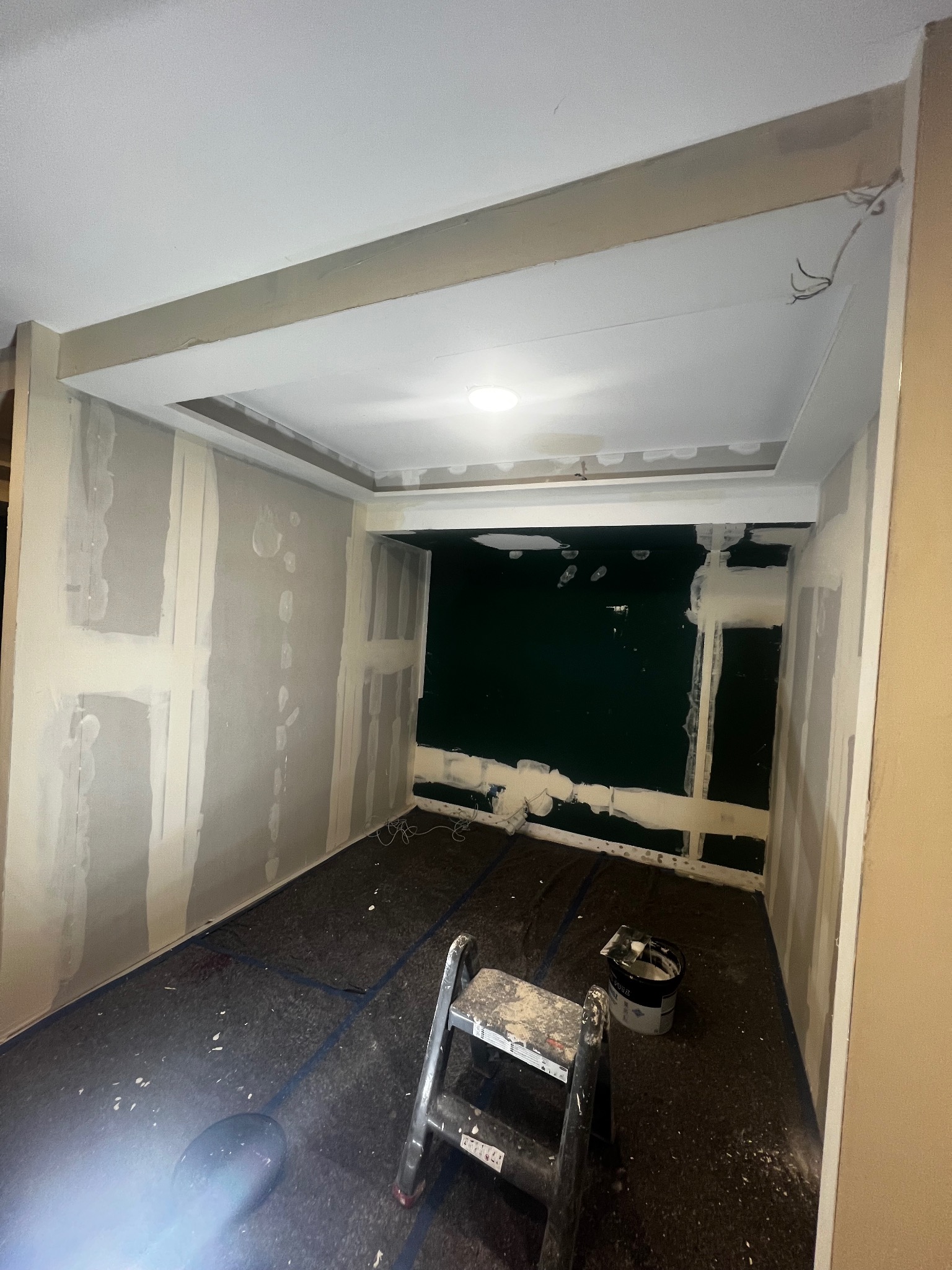 Pokój w trakcie prac wykończeniowych: ściany pokryte warstwą gładzi, widoczna drabina malarska i wiadro z zaprawą na zabezpieczonej podłodze, sufit z elementami płyty karton-gips i wystającymi...