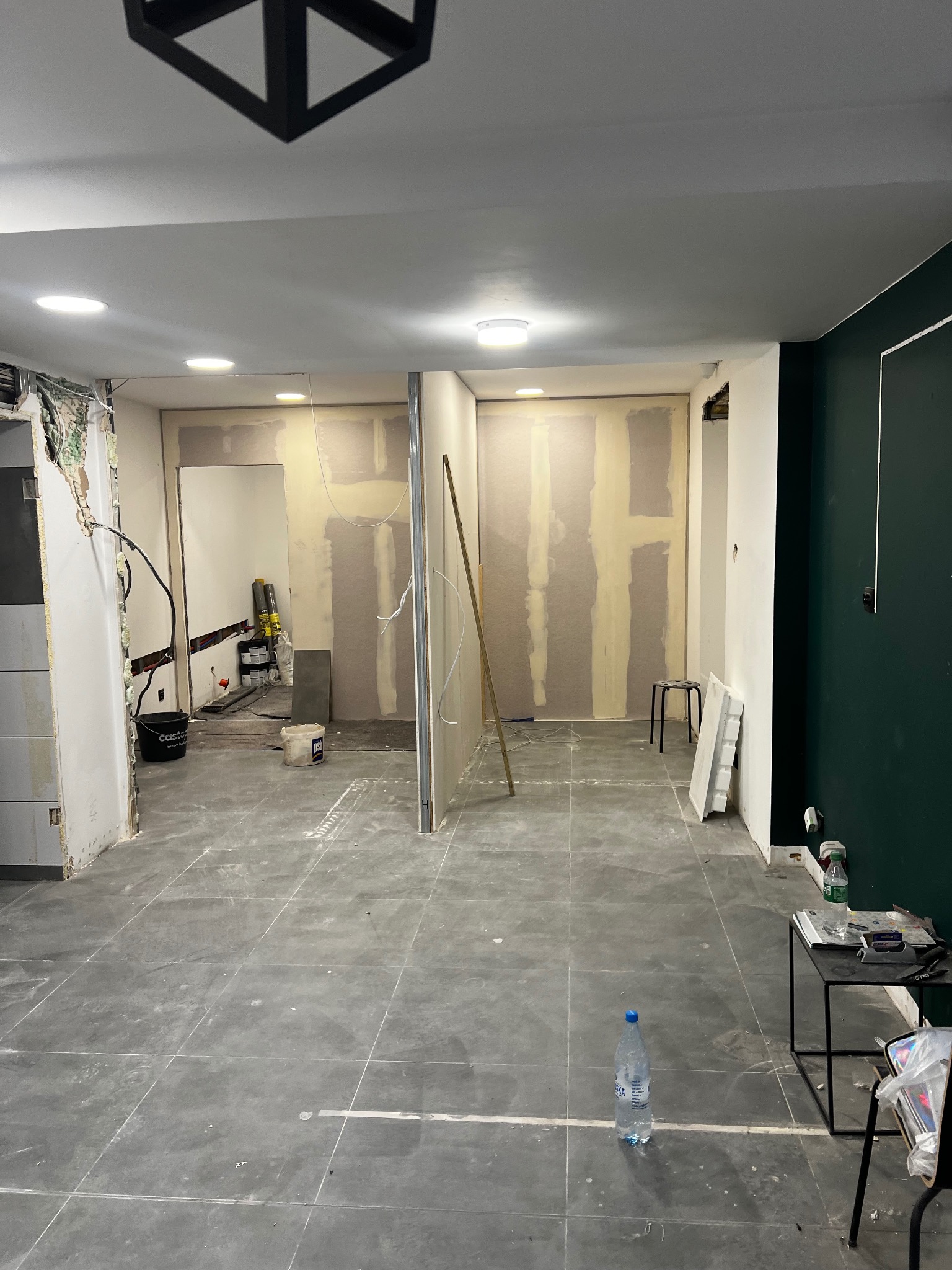 Wnętrze w trakcie prac wykończeniowych: ściany z częściowo nałożoną gładzią, płyty karton-gips, widoczne instalacje elektryczne, szare płytki podłogowe i butelka wody mineralnej na podłodze.