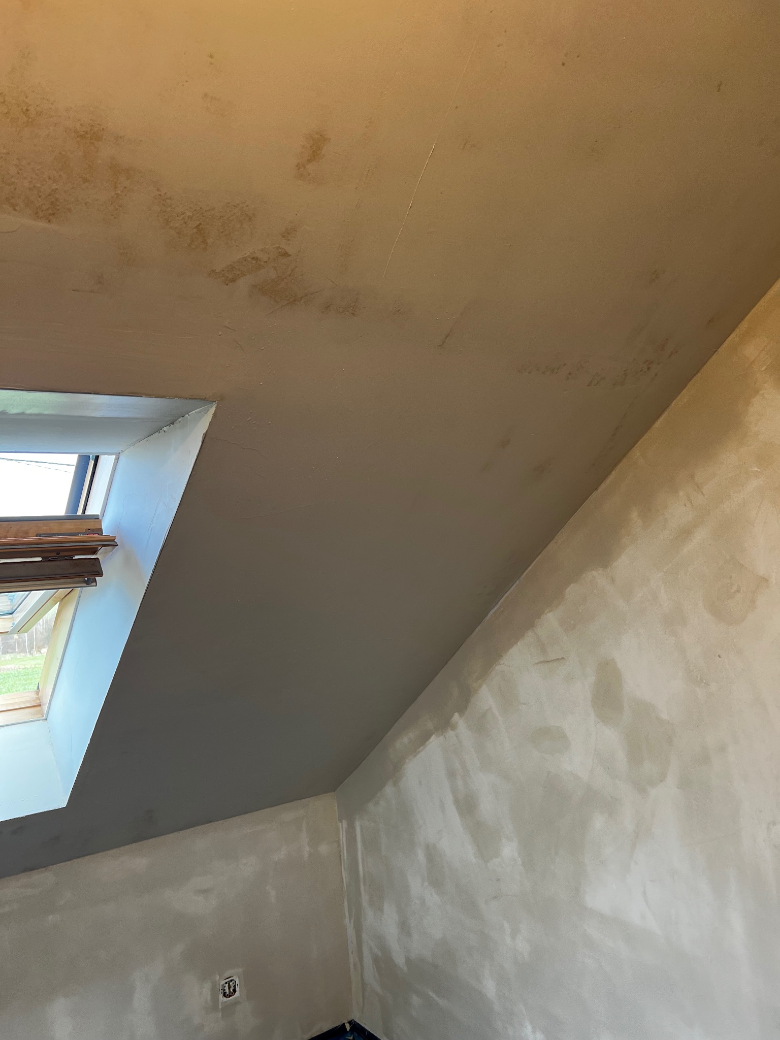 Skośny pokój na poddaszu w trakcie gładzenia ścian i sufitu, widoczne okno dachowe i niedoskonałości na powierzchni. Ściany częściowo pokryte gładzią.