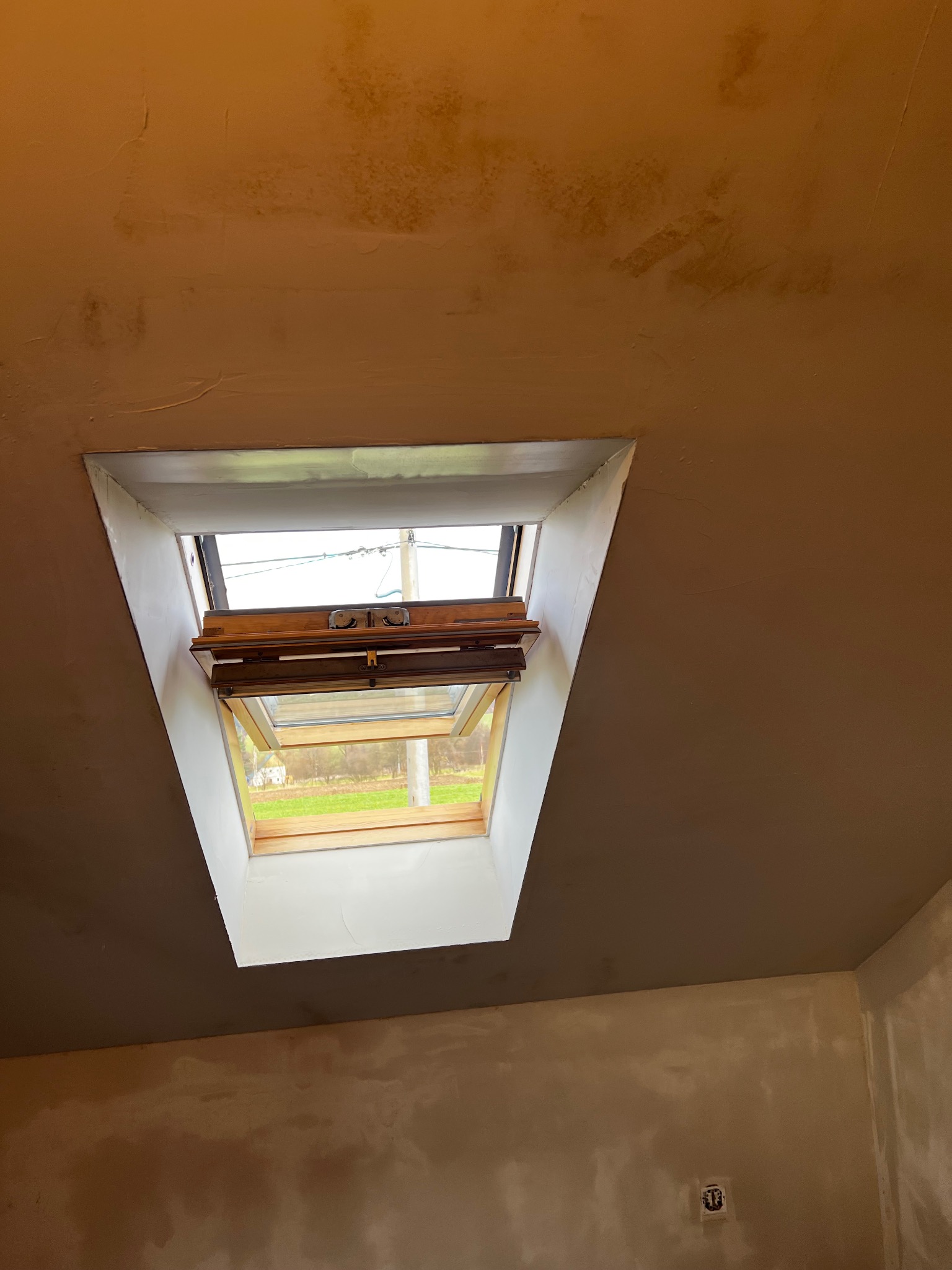 Ściana i sufit po nałożeniu gładzi szpachlowej, widoczne okno dachowe z drewnianą ramą. Ślady po szpachlowaniu na suficie i ścianach. W tle widok na zewnątrz przez otwarte okno.