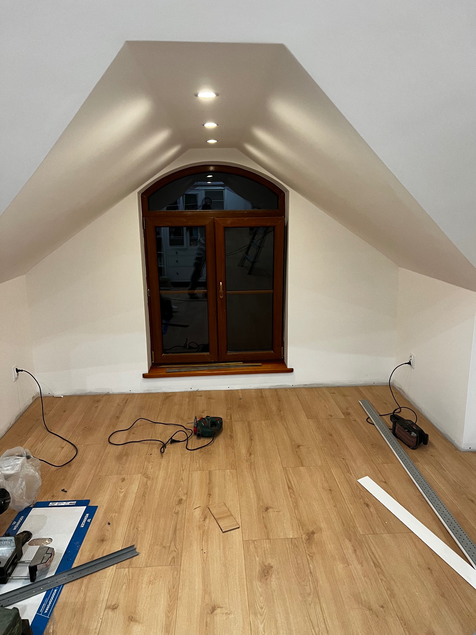 Układanie paneli podłogowych w pokoju na poddaszu z oknem w drewnianej ramie. Widoczne narzędzia i materiały montażowe. Oświetlenie punktowe w suficie.