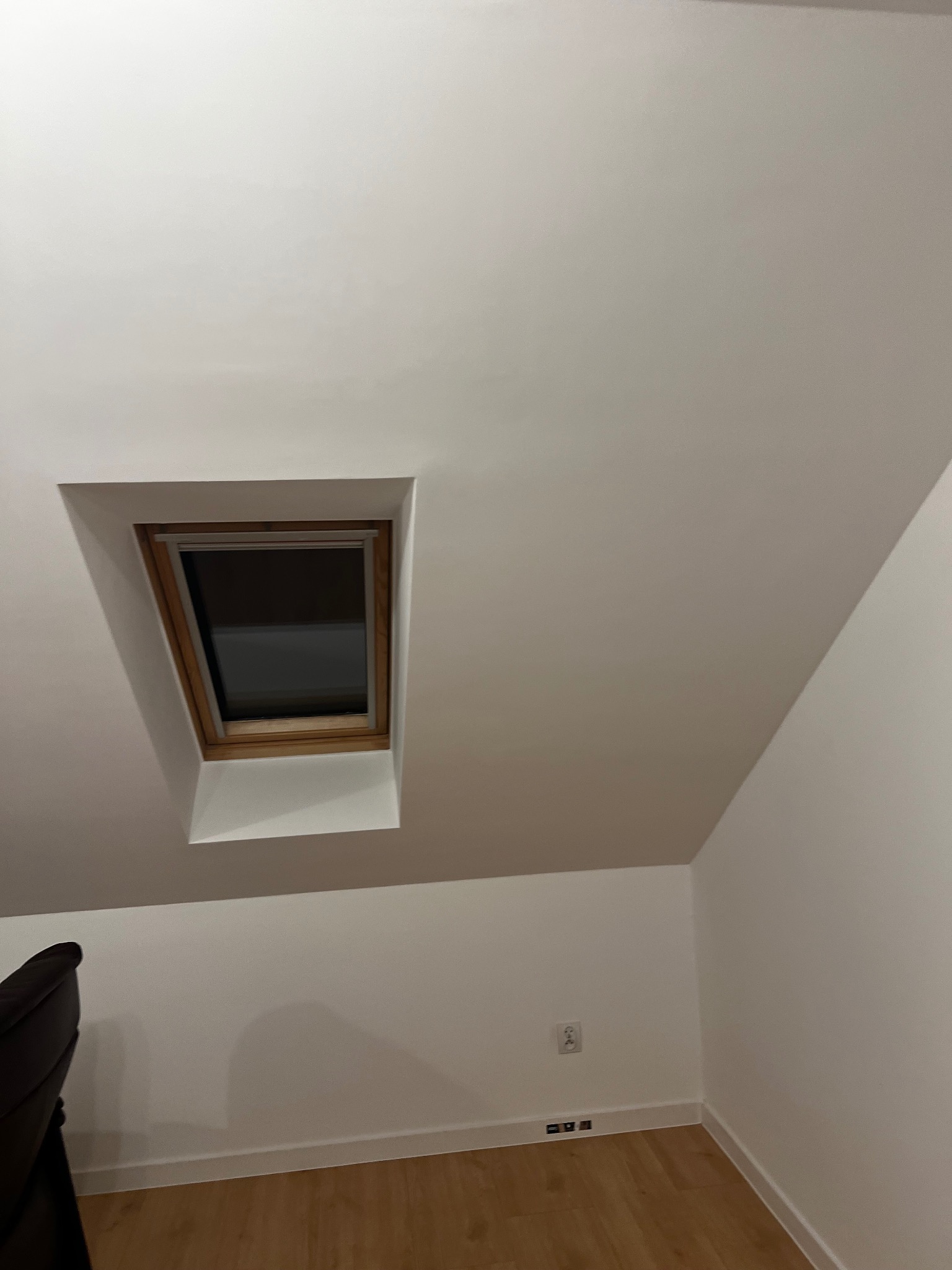 Białe poddasze z oknem dachowym w drewnianej ramie. Widoczne gładkie ściany, drewniana podłoga i fragment mebla. Minimalistyczne wykończenie wnętrza.