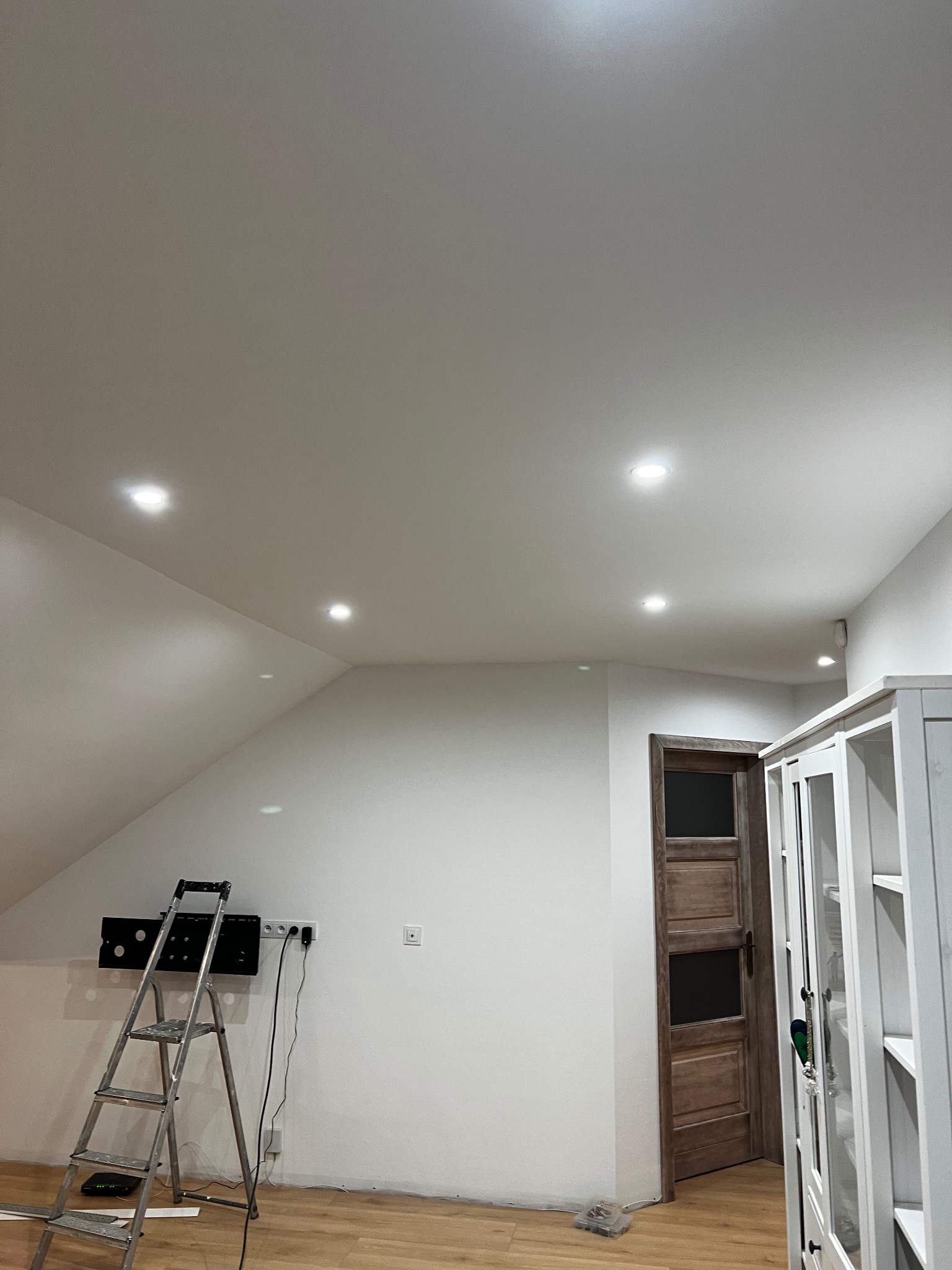 Białe wnętrze poddasza z zamontowanymi punktowymi lampami sufitowymi. Widoczna drabina, szafa i drewniane drzwi. Świeżo wykończone ściany.