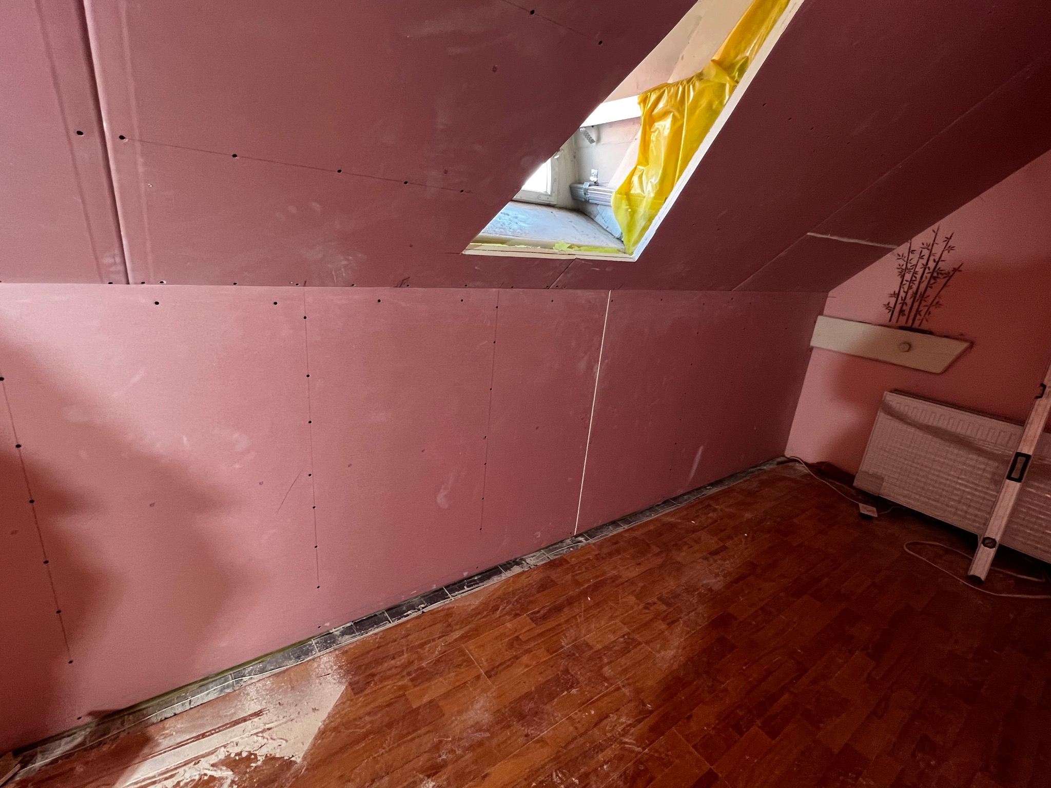 Poddasze z oknem dachowym w trakcie wykańczania płytami gipsowo-kartonowymi, widoczne ślady po szpachlowaniu, drewniana podłoga i grzejnik.