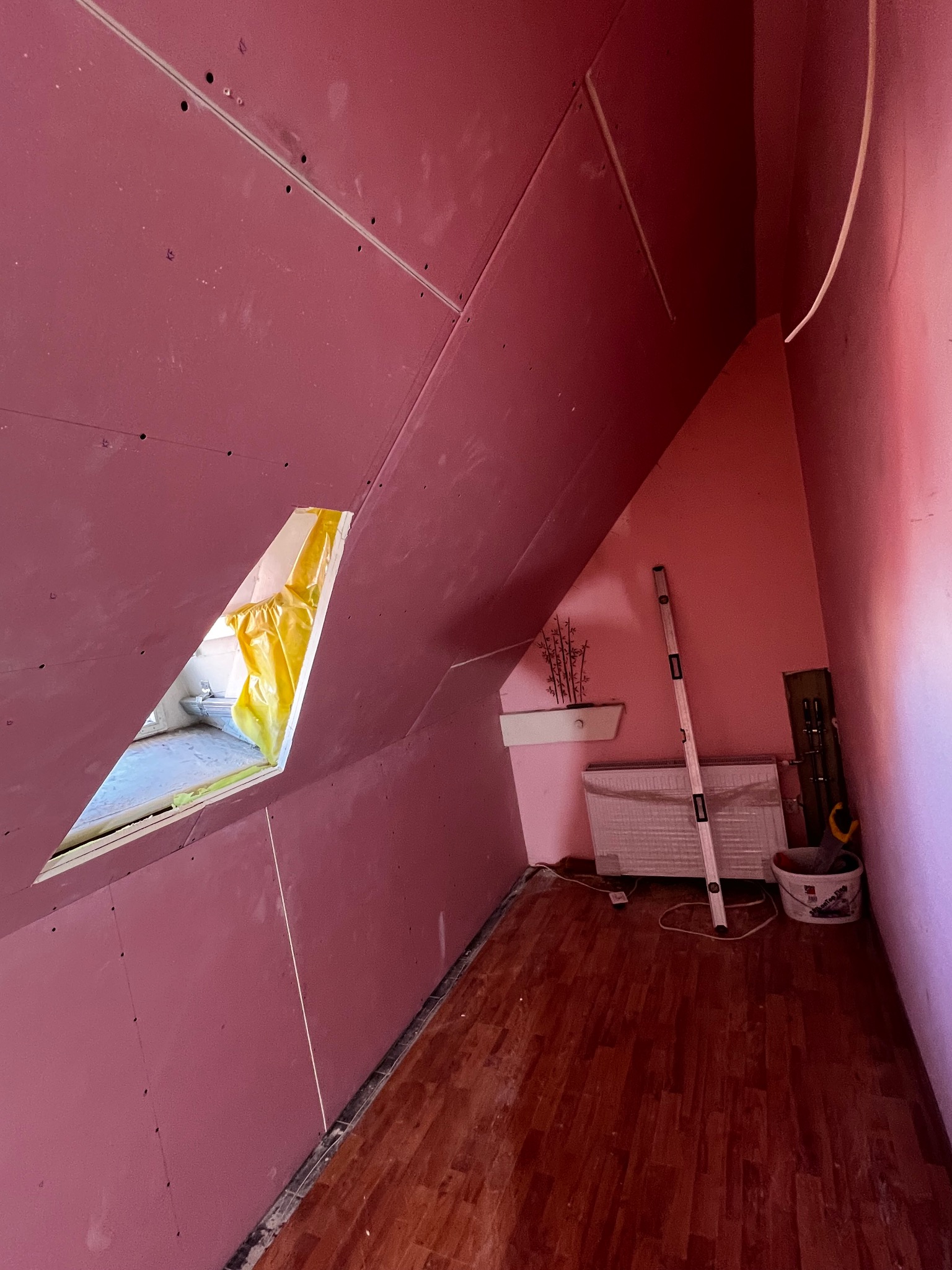 Poddasze z oknem dachowym w trakcie wykańczania płytami kartonowo-gipsowymi w kolorze różowym. Widoczne narzędzia i poziomica. Parkiet na podłodze.