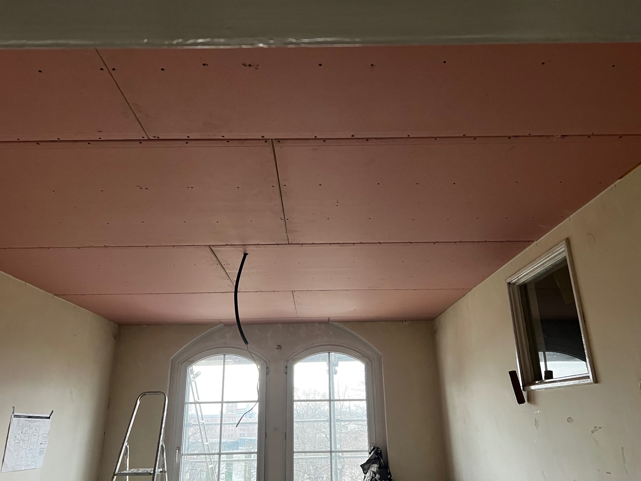 Różowe płyty karton-gips na suficie w pokoju z oknami o łukowatych ramach. Widoczna drabina i zwisający kabel. Etap prac wykończeniowych.