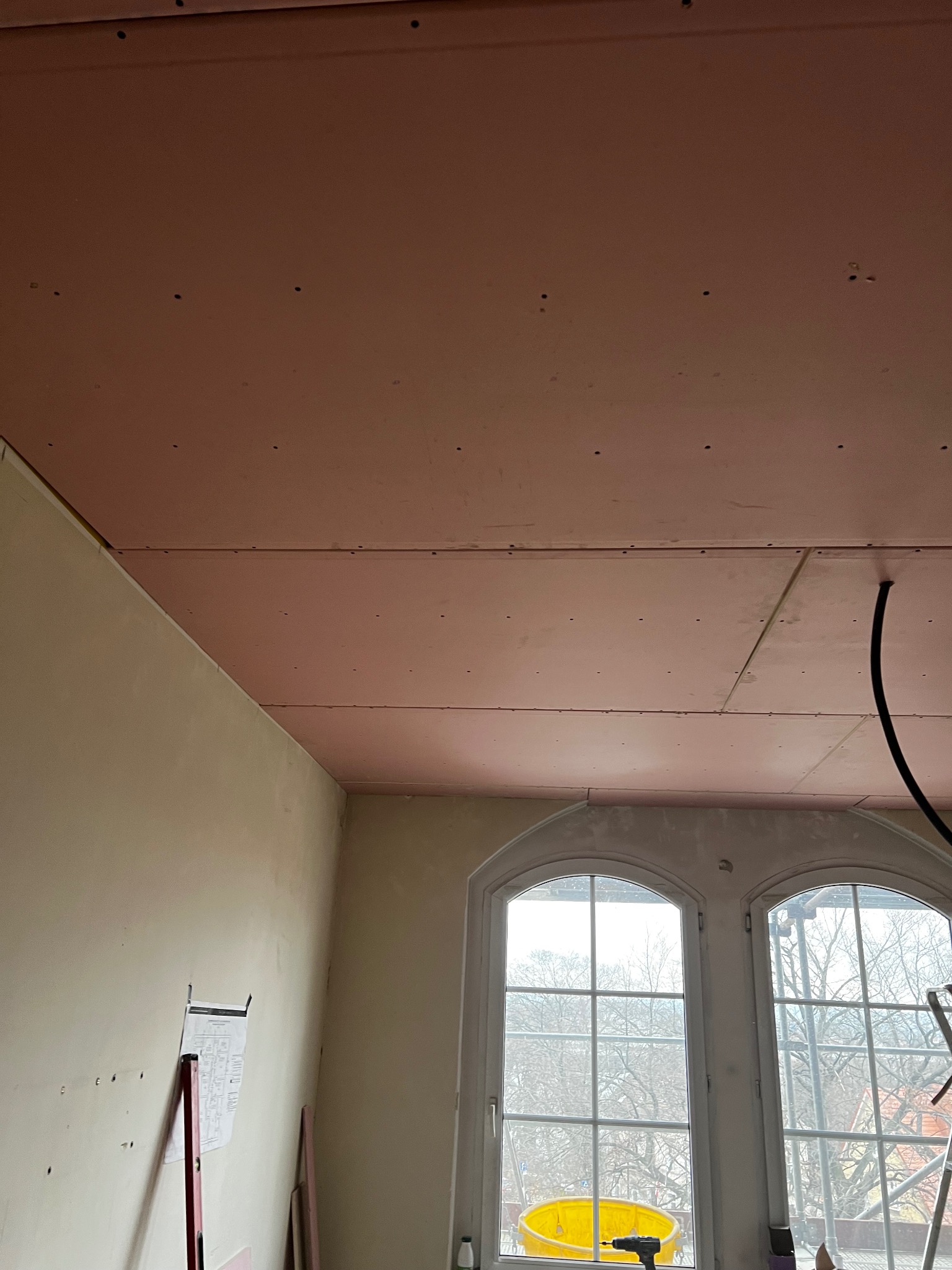 Różowe płyty karton-gips na suficie, mocowane wkrętami, widok z dołu. W tle okno z podwójnymi łukami i drabina. Na ścianie plan pomieszczenia.