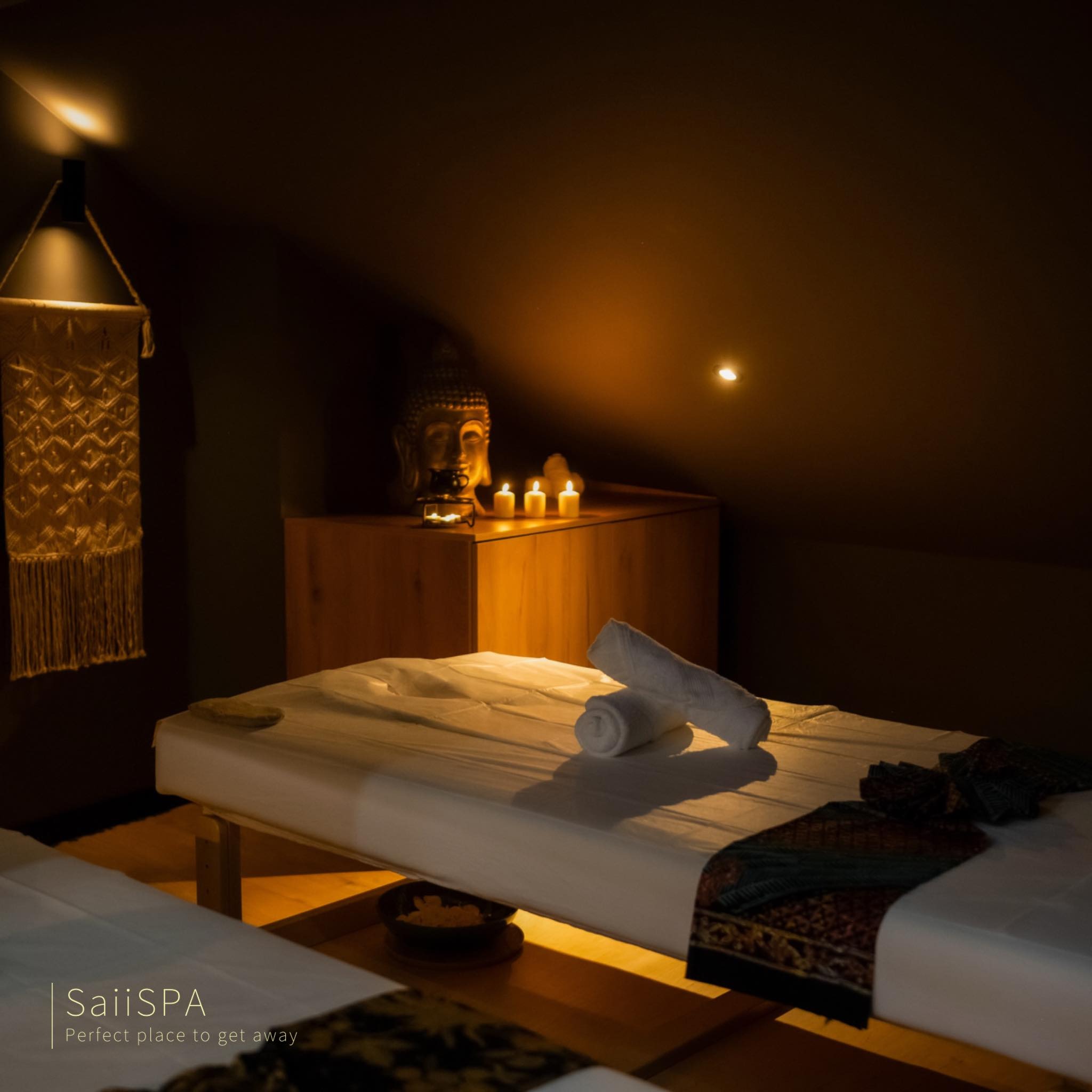 Dwa łóżka do masażu w ciemnym pomieszczeniu SPA, z ręcznikami i dekoracyjnym bieżnikiem, oświetlone świecami i lampą, z drewnianą szafką zwieńczoną figurką Buddy w tle.