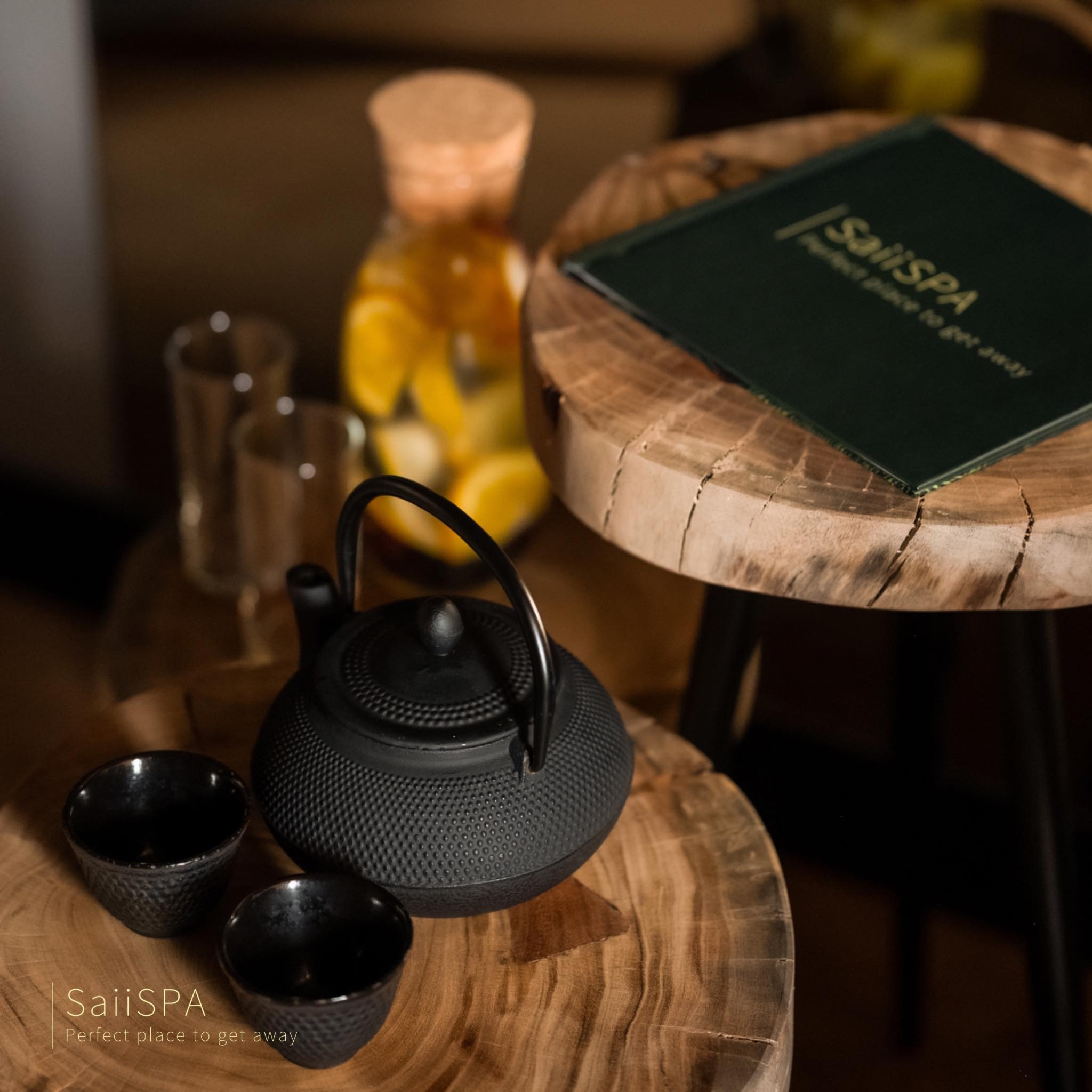 Czarny żeliwny czajnik i dwie czarki na drewnianym stoliku, w tle butelka z wodą i owocami oraz menu z logo SaiiSPA, tworzące relaksującą atmosferę.