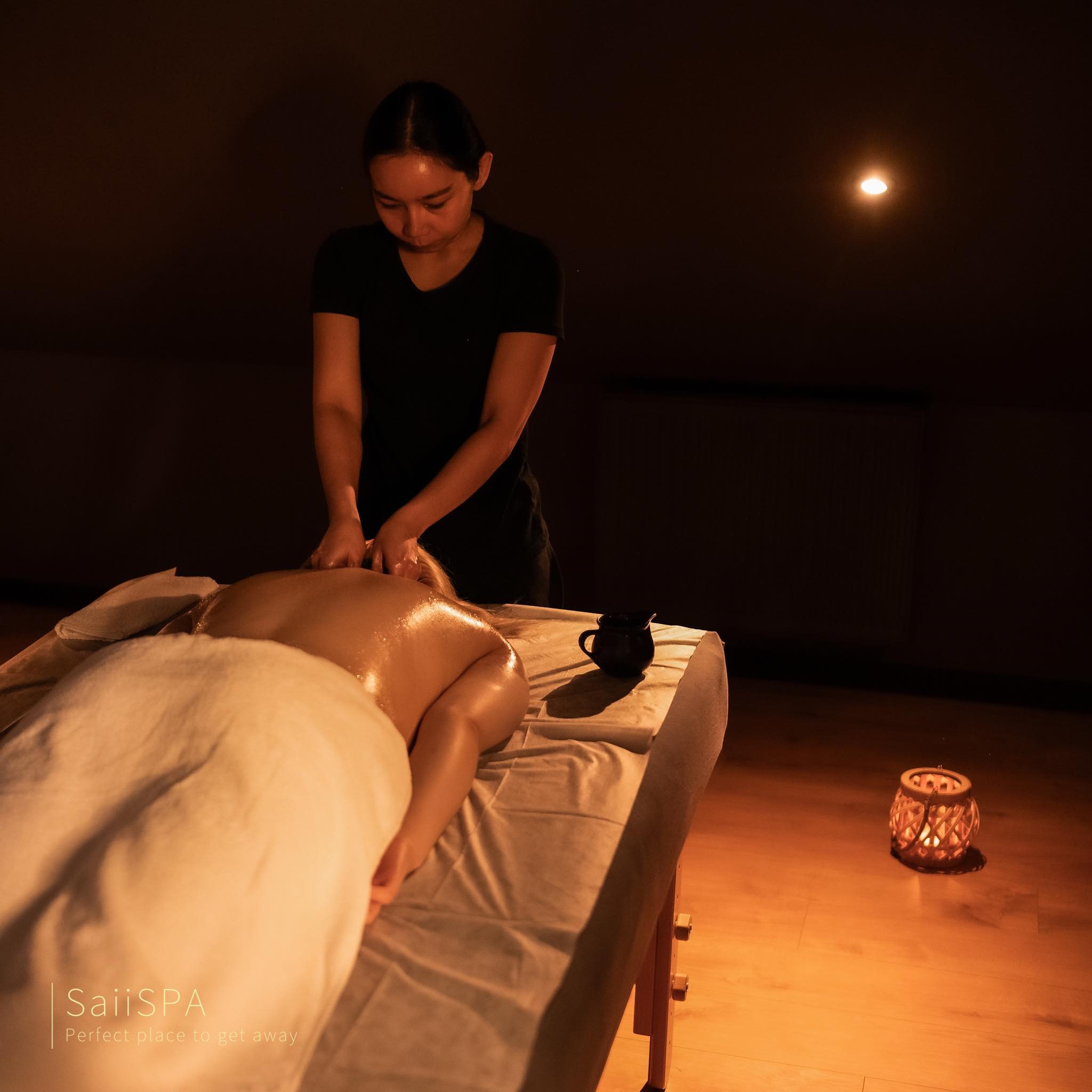 Kobieta masuje plecy drugiej osobie leżącej na stole w ciemnym pomieszczeniu oświetlonym świecą i lampą, obok stoi dzbanek.