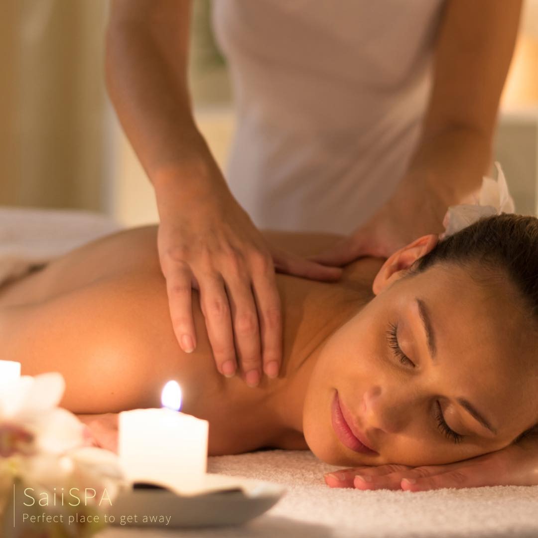 Kobieta relaksuje się podczas masażu pleców w spa, przy blasku świec i dekoracji kwiatowych, z widocznym logo 'SaiiSPA' w lewym dolnym rogu.