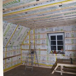 Usługi ogolnobudowlane - Poddasze w trakcie ocieplania, widoczne drewniane belki, folia paroizolacyjna z napisem INTELLO PLUS, drabina aluminiowa i okno dachowe.