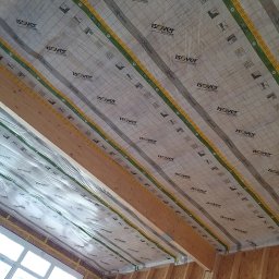 Usługi ogolnobudowlane - Ocieplenie poddasza folią paroizolacyjną Isover Vario XtraSafe, widok na drewnianą konstrukcję dachu i okno w Koszalinie.