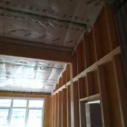 Wnętrze w trakcie ocieplania poddasza: widoczne drewniane belki konstrukcyjne, folia izolacyjna Isover na suficie i ścianach, okno z żółtą taśmą uszczelniającą.