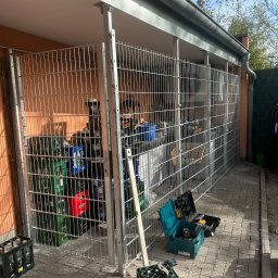 Usługi ogolnobudowlane - Konstrukcja z metalowej siatki tworząca zadaszone miejsce do składowania, widoczne skrzynki z butelkami, narzędzia w kufrach na kostce brukowej.