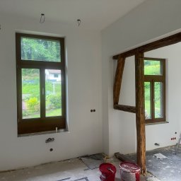 Usługi ogolnobudowlane - Wnętrze w trakcie prac wykończeniowych: pomalowane ściany na biało, drewniane ramy okienne, widoczne puszki elektryczne, fragment drewnianej konstrukcji, drabina i puszki z farbą na podłodze.