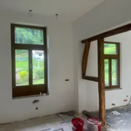 Wnętrze w trakcie prac wykończeniowych: pomalowane ściany na biało, drewniane ramy okienne, widoczne puszki elektryczne, fragment drewnianej konstrukcji, drabina i puszki z farbą na podłodze.