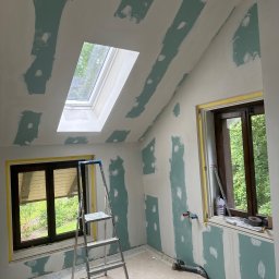 Usługi ogolnobudowlane - Poddasze w trakcie szpachlowania, widoczne pasy gipsu na ścianach i suficie skośnym, okno dachowe, drabina aluminiowa, okna zabezpieczone folią, narzędzia malarskie i wiadro na podłodze.