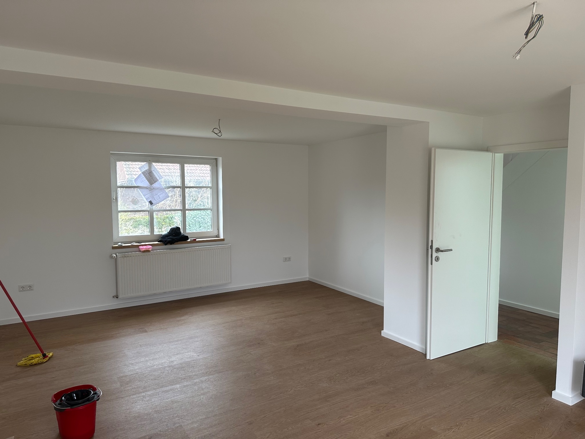 Pusta, jasna przestrzeń z panelami podłogowymi, świeżo pomalowanymi ścianami i białymi drzwiami, z mopem i wiadrem w rogu, oraz planami na oknie.