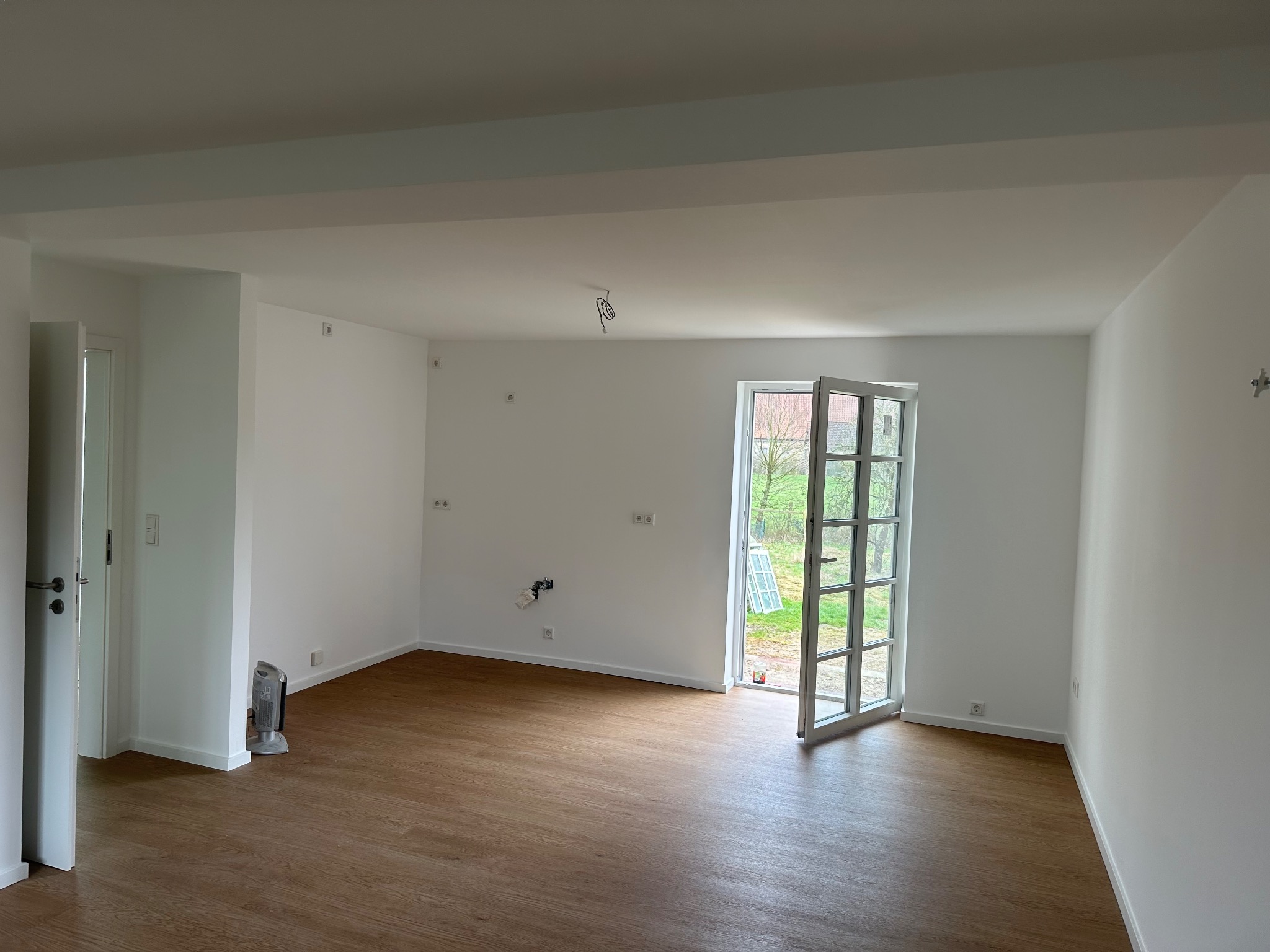 Pusty, świeżo wykończony pokój z drewnianą podłogą, białymi ścianami i drzwiami balkonowymi uchylonymi na zewnątrz, widoczne wystające kable elektryczne i grzejnik elektryczny.
