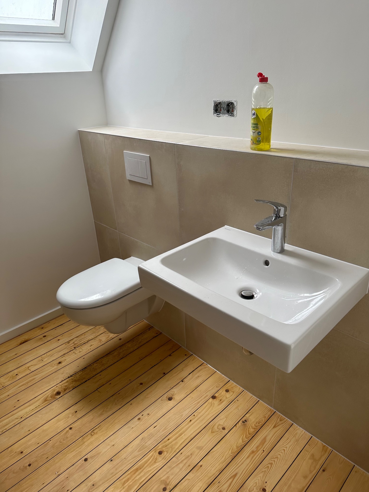 Nowoczesna łazienka na poddaszu z drewnianą podłogą, wiszącą toaletą i umywalką, widoczne niedokończone gniazdka elektryczne i butelka płynu do mycia na półce.