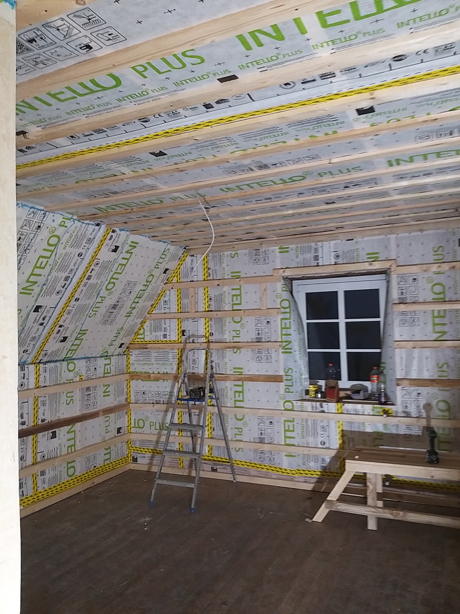 Poddasze w trakcie ocieplania, widoczne drewniane belki, folia paroizolacyjna z napisem INTELLO PLUS, drabina aluminiowa i okno dachowe.