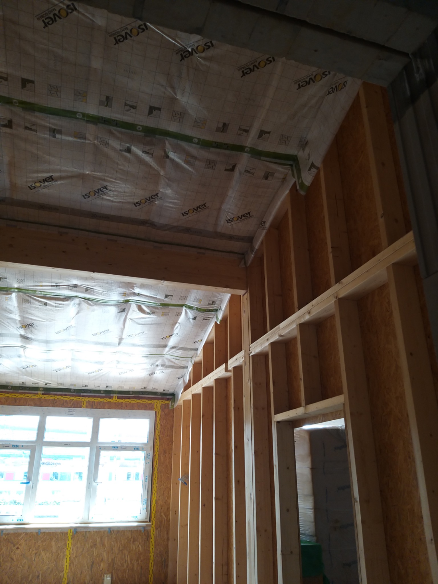 Wnętrze w trakcie ocieplania poddasza: widoczne drewniane belki konstrukcyjne, folia izolacyjna Isover na suficie i ścianach, okno z żółtą taśmą uszczelniającą.