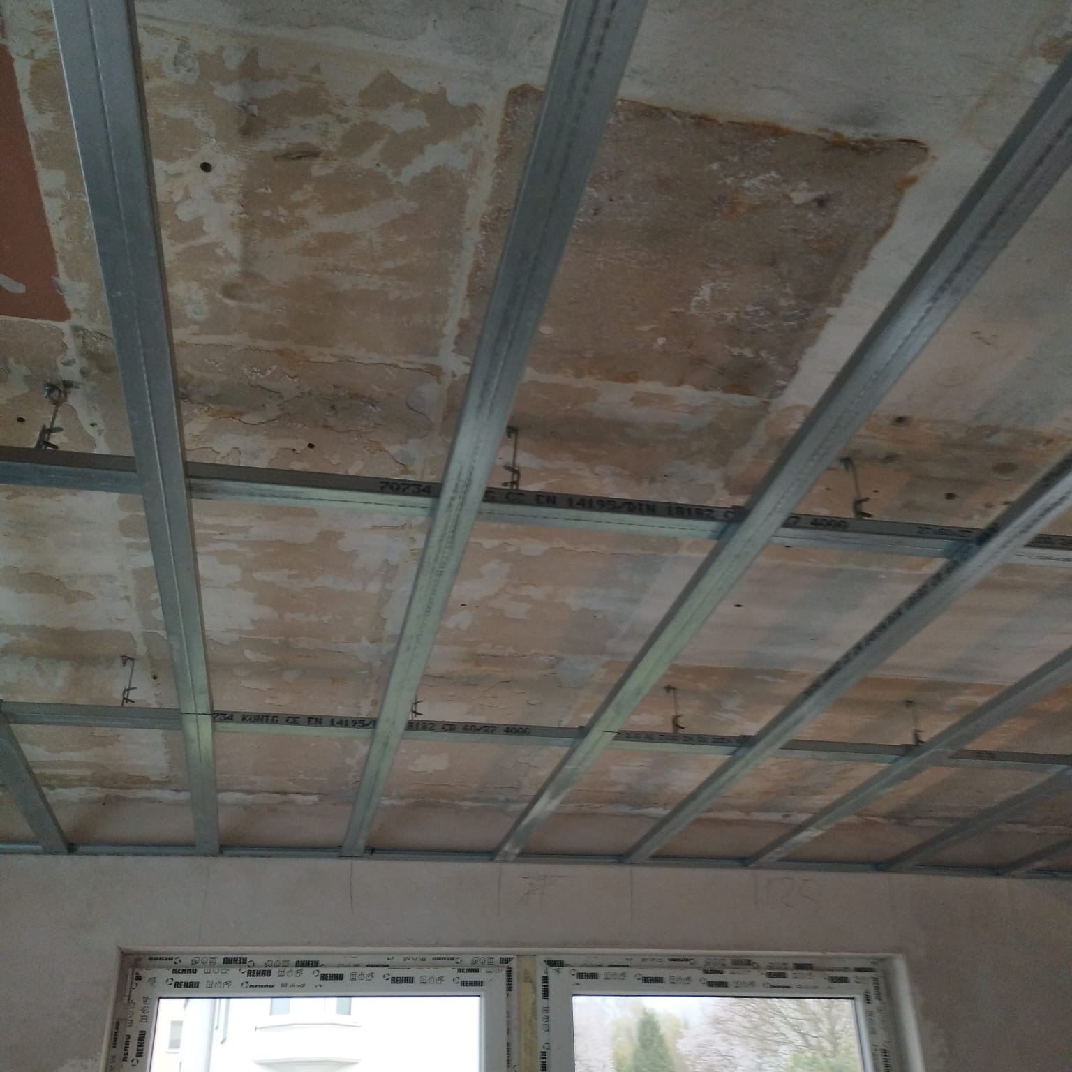 Stelaż z profili metalowych zamontowany pod nierównym, starym stropem przygotowany do montażu płyt gipsowo-kartonowych, widok z dołu na tle okna.