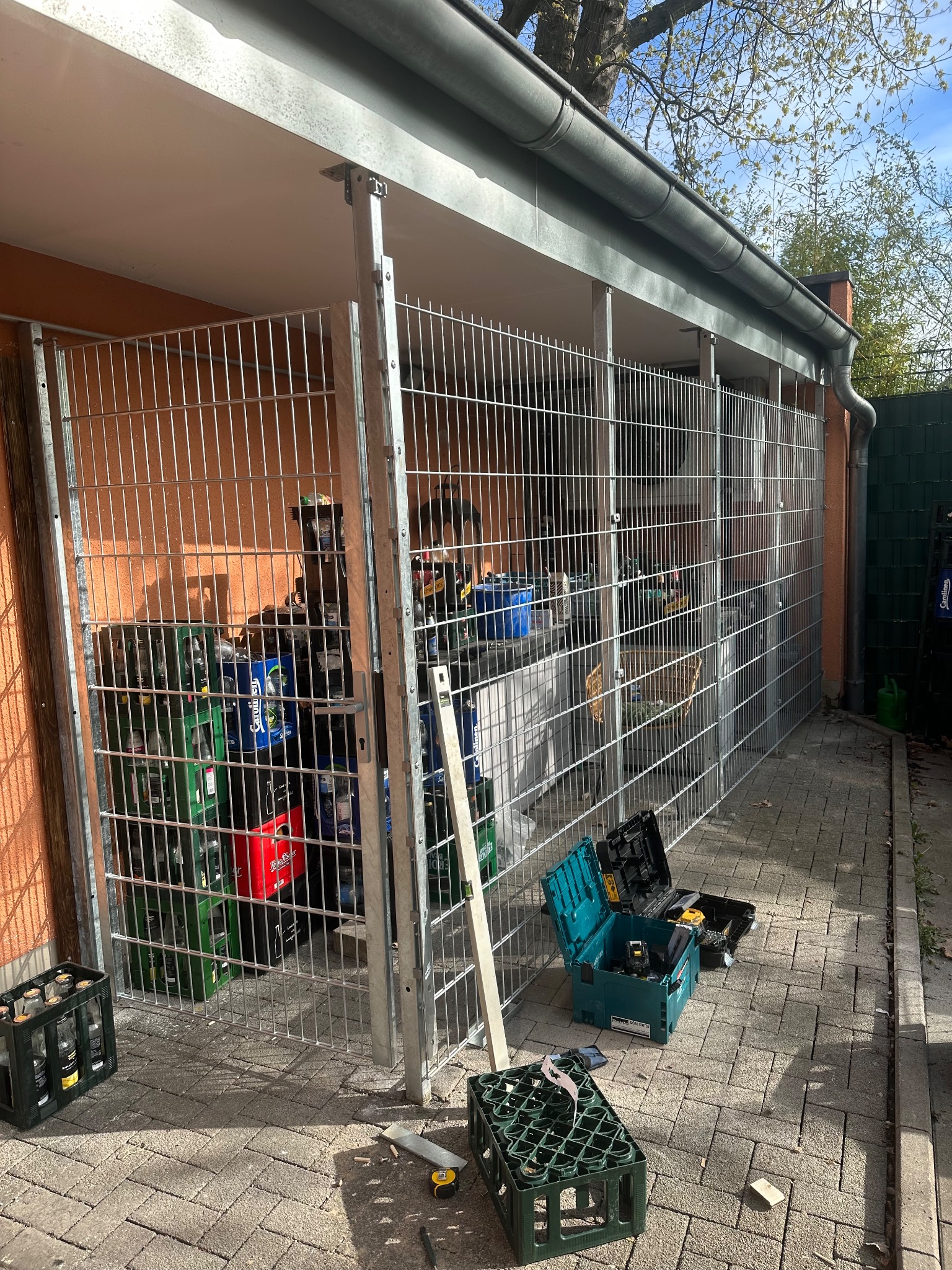 Konstrukcja z metalowej siatki tworząca zadaszone miejsce do składowania, widoczne skrzynki z butelkami, narzędzia w kufrach na kostce brukowej.