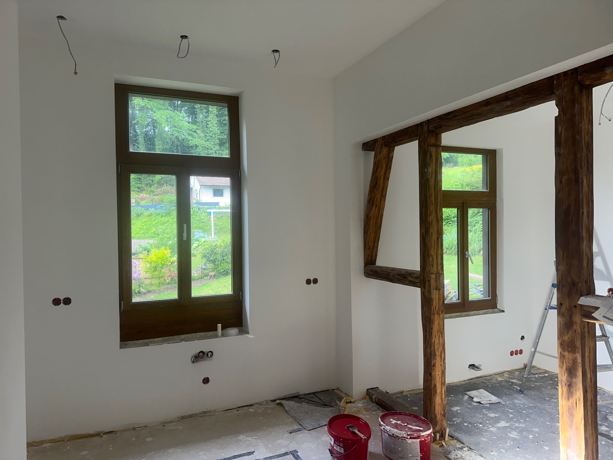 Wnętrze w trakcie prac wykończeniowych: pomalowane ściany na biało, drewniane ramy okienne, widoczne puszki elektryczne, fragment drewnianej konstrukcji, drabina i puszki z farbą na podłodze.