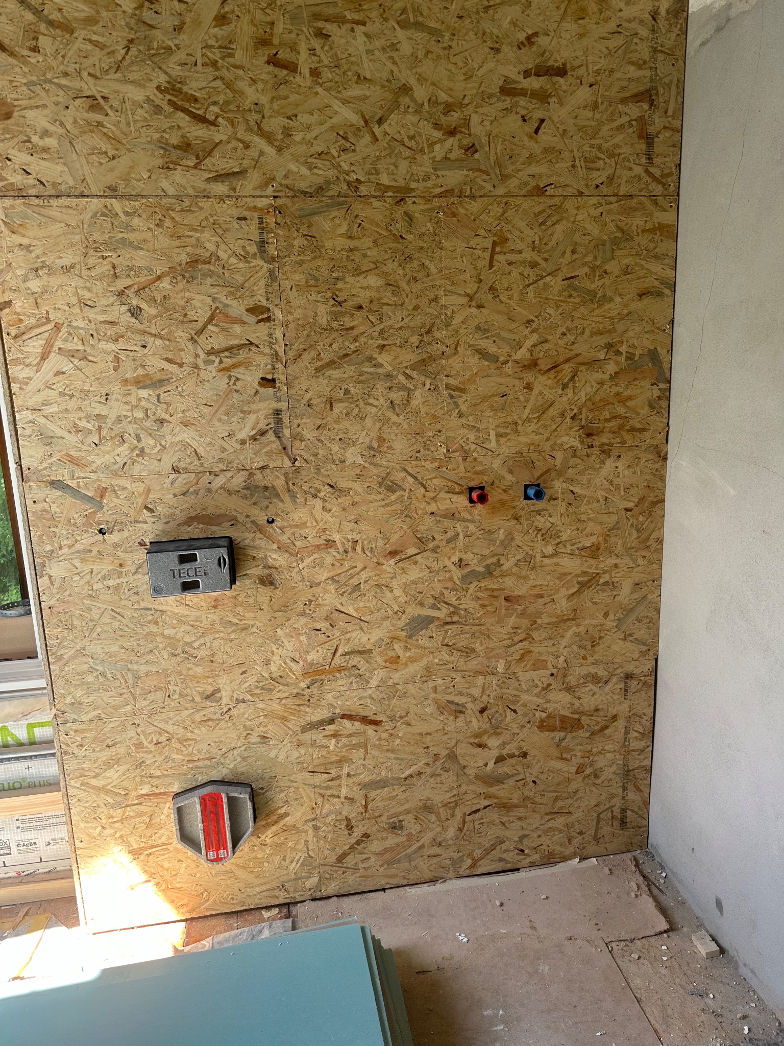 Ściana z płyty OSB z zamontowanymi elementami instalacji wodnej (czerwony i niebieski otwór) oraz stelażem podtynkowym TECE, widoczne płyty karton gips na podłodze.