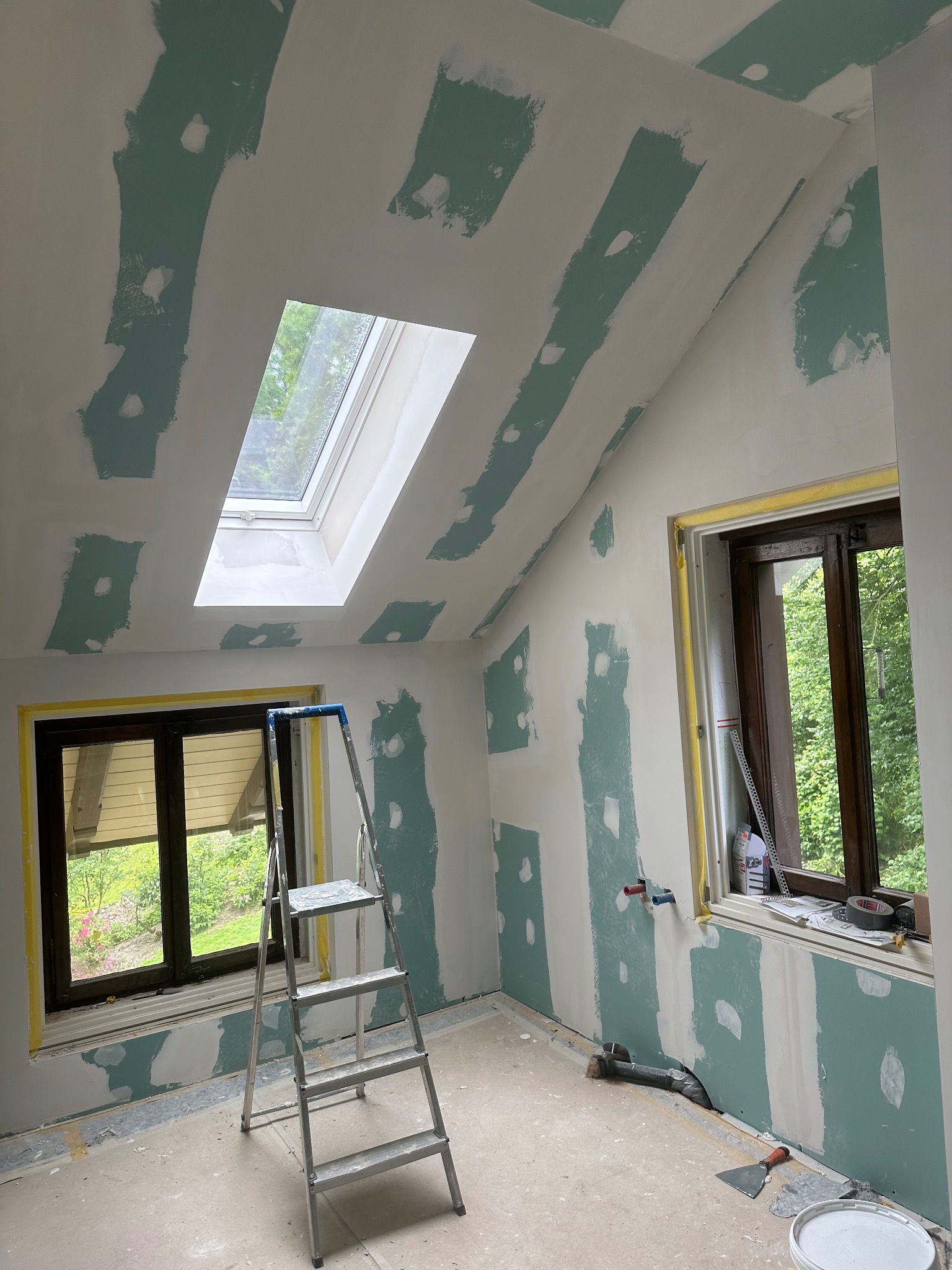 Poddasze w trakcie szpachlowania, widoczne pasy gipsu na ścianach i suficie skośnym, okno dachowe, drabina aluminiowa, okna zabezpieczone folią, narzędzia malarskie i wiadro na podłodze.