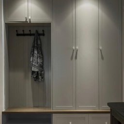 PKN Estate Sp. z o.o. - Szara szafa wnękowa z drewnianym siedziskiem i wieszakami, oświetlona punktowo, na tle drewnianej podłogi, z fragmentem ciemnego stołu po prawej stronie.